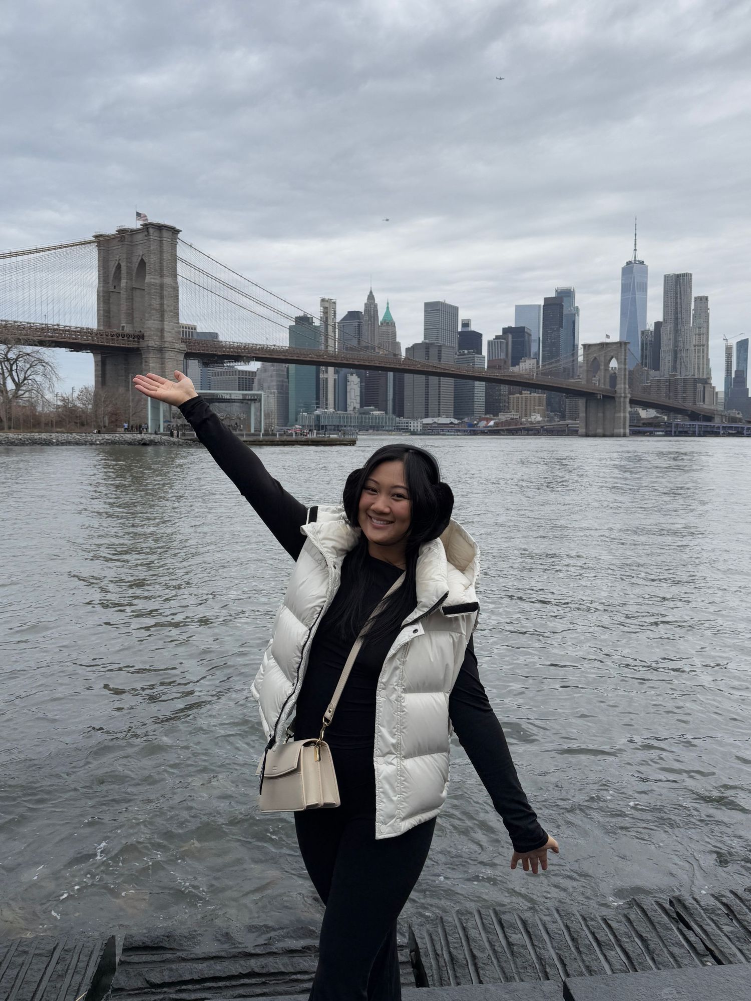 Allyson posing joyfully in front of Brooklyn Bridge and Manhattan skyline on cloudy day.