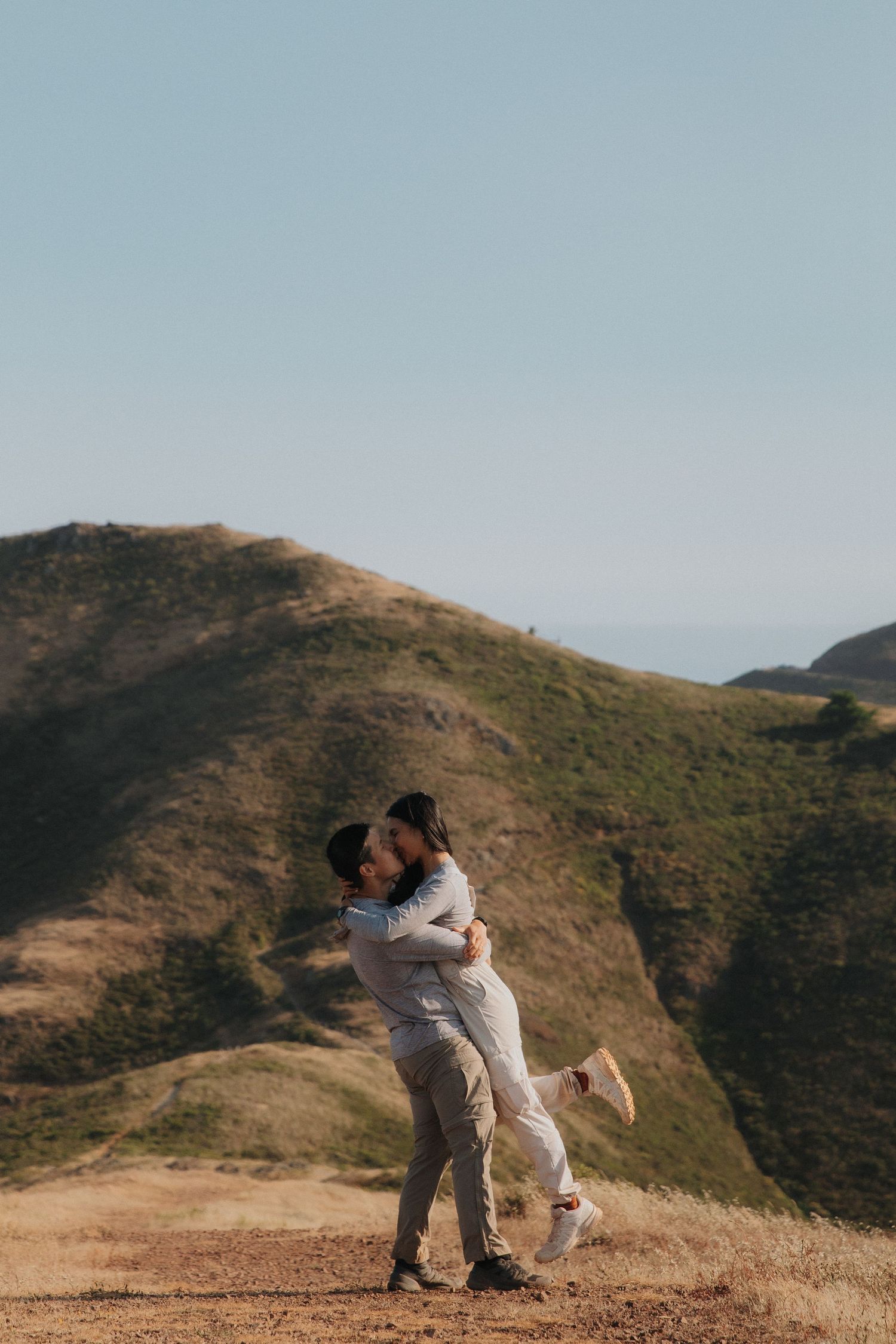 Two people embrace warmly while standing on a grassy hilltop overlooking rolling hills and mountainous terrain.