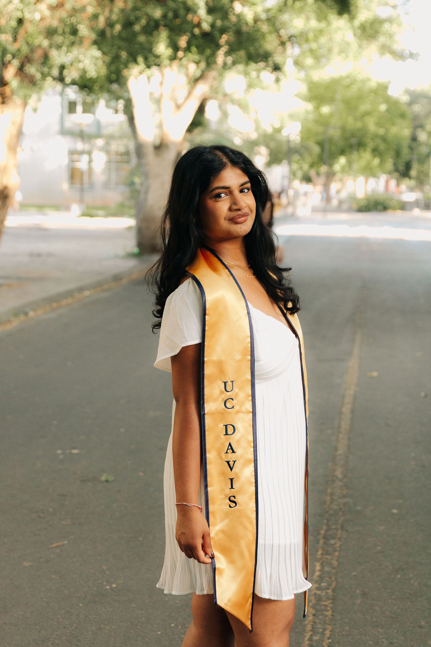 Graduate wearing white dress and gold UC Davis graduation sash poses on tree-lined street during golden hour.