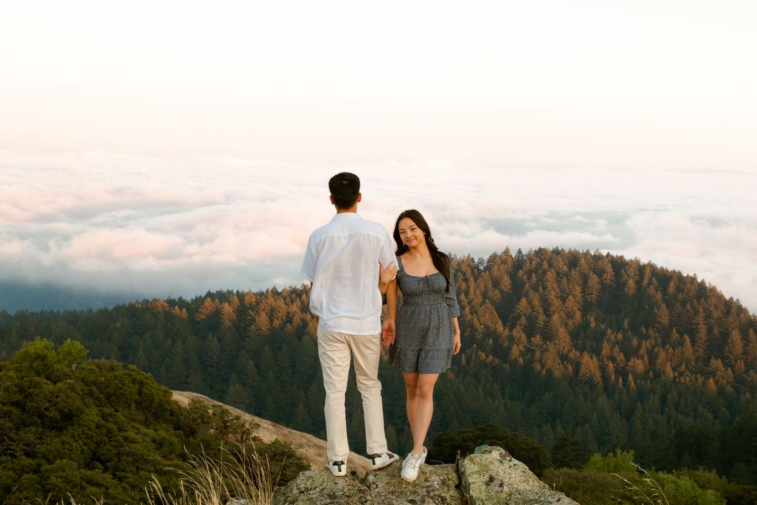 A couple stands on a mountain summit overlooking a forested landscape during golden hour.