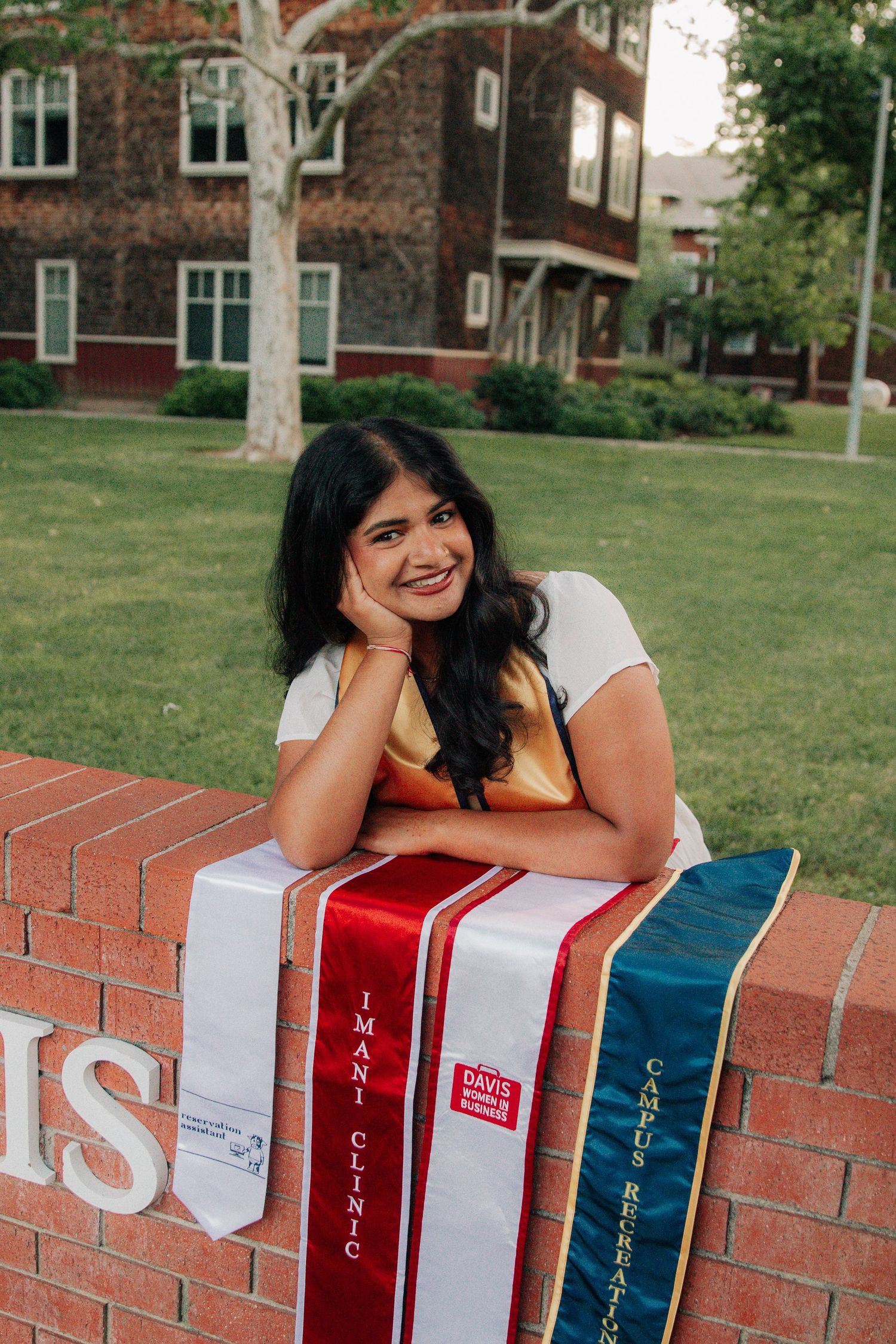 A person sits smiling beside large decorative books on a brick wall at a university campus on a sunny day.