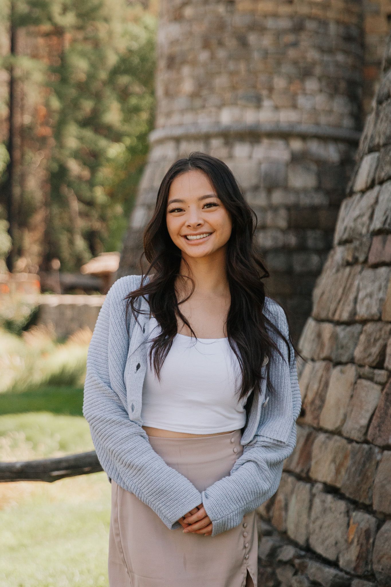 A woman wearing a gray cardigan poses against a stone wall in a casual outdoor portrait.