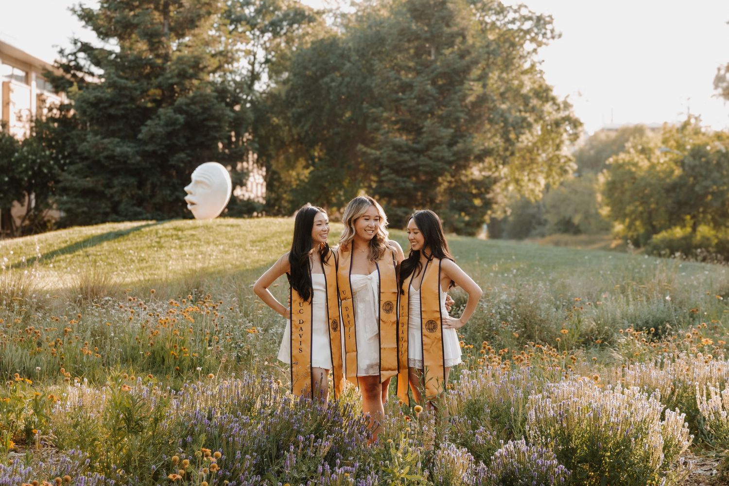 Group dressed in flowy, cream-colored dresses walks through a sunlit meadow during golden hour.