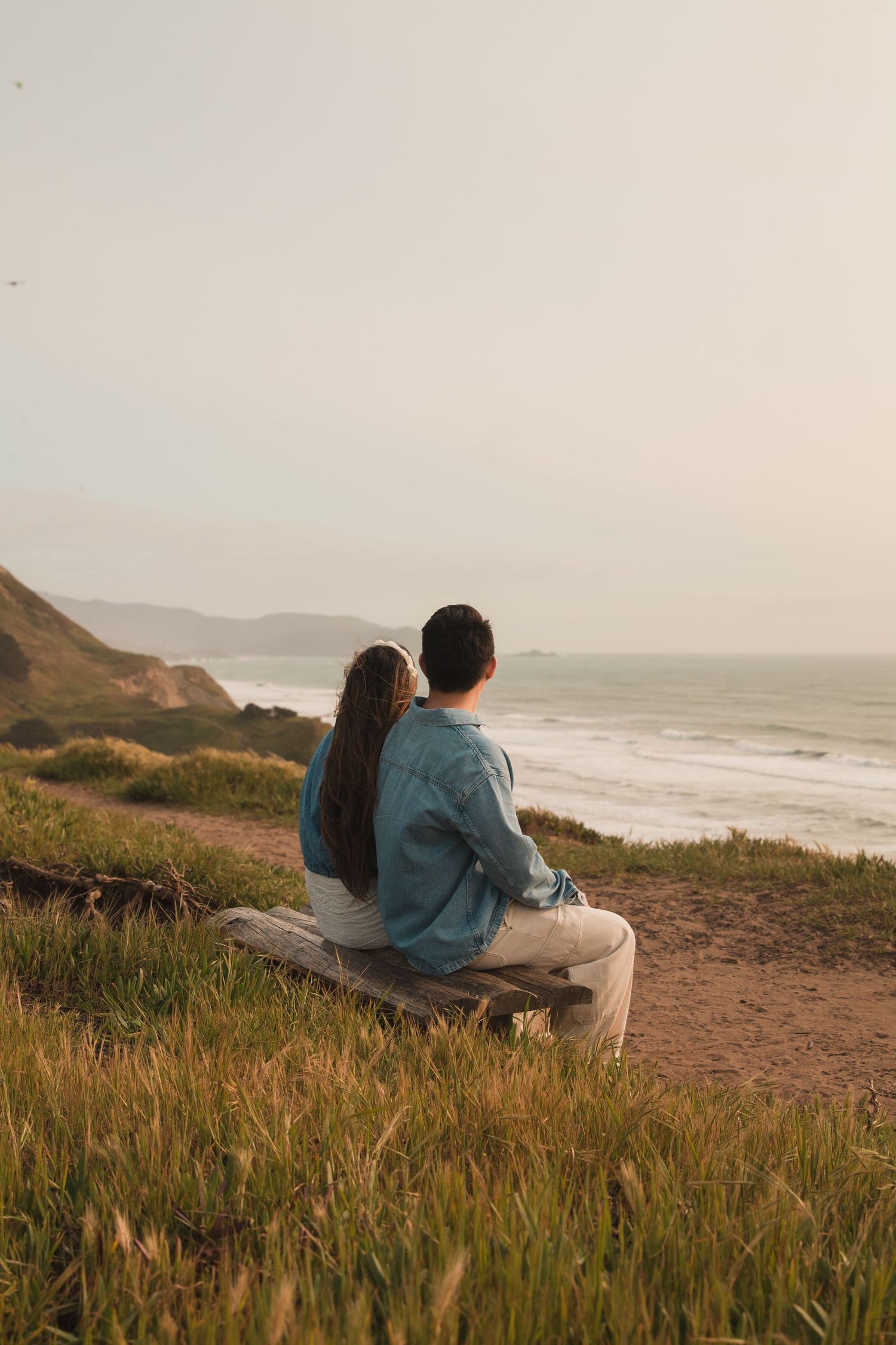 Couple sits on clifftop bench overlooking foggy coastal view.