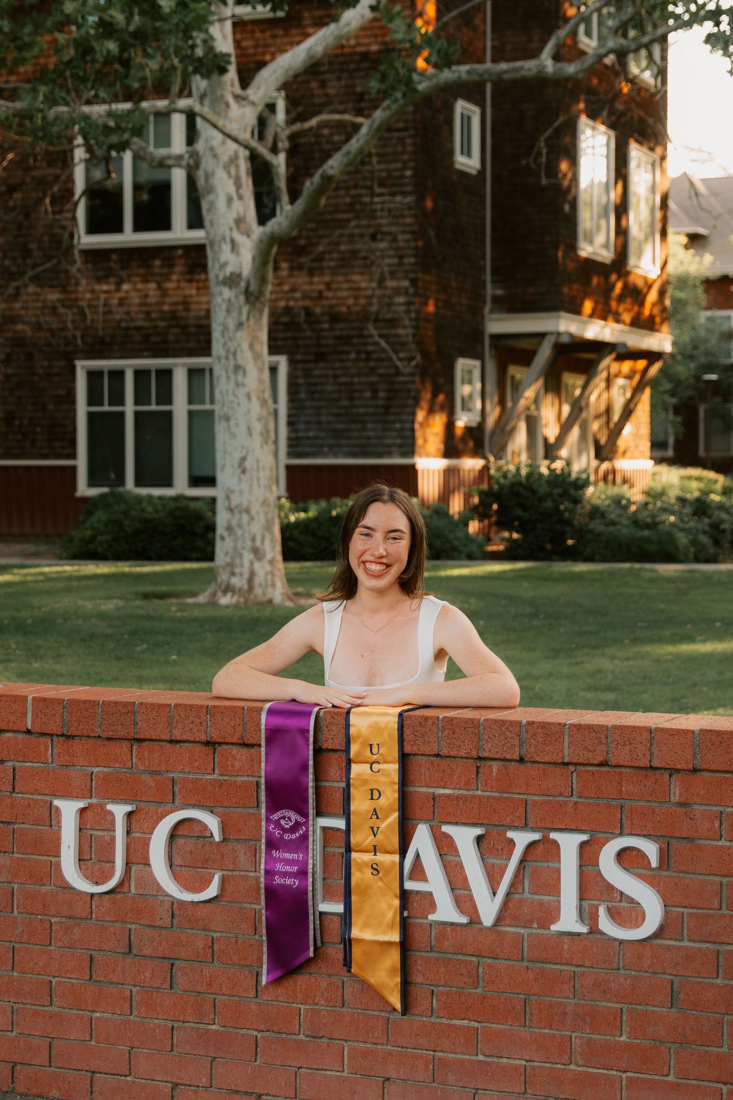 A graduate poses with purple and gold graduation stoles at the UC Davis brick entrance sign on a sunny day.