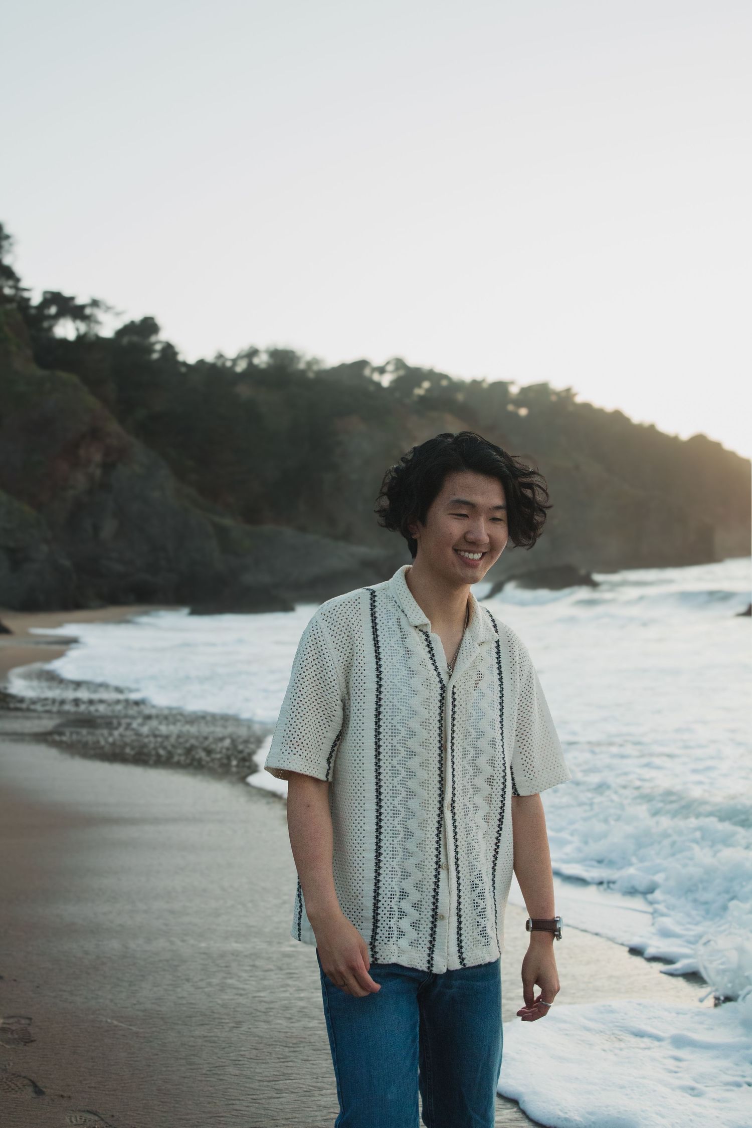 Person in patterned shirt smiling on coastal beach at sunset.