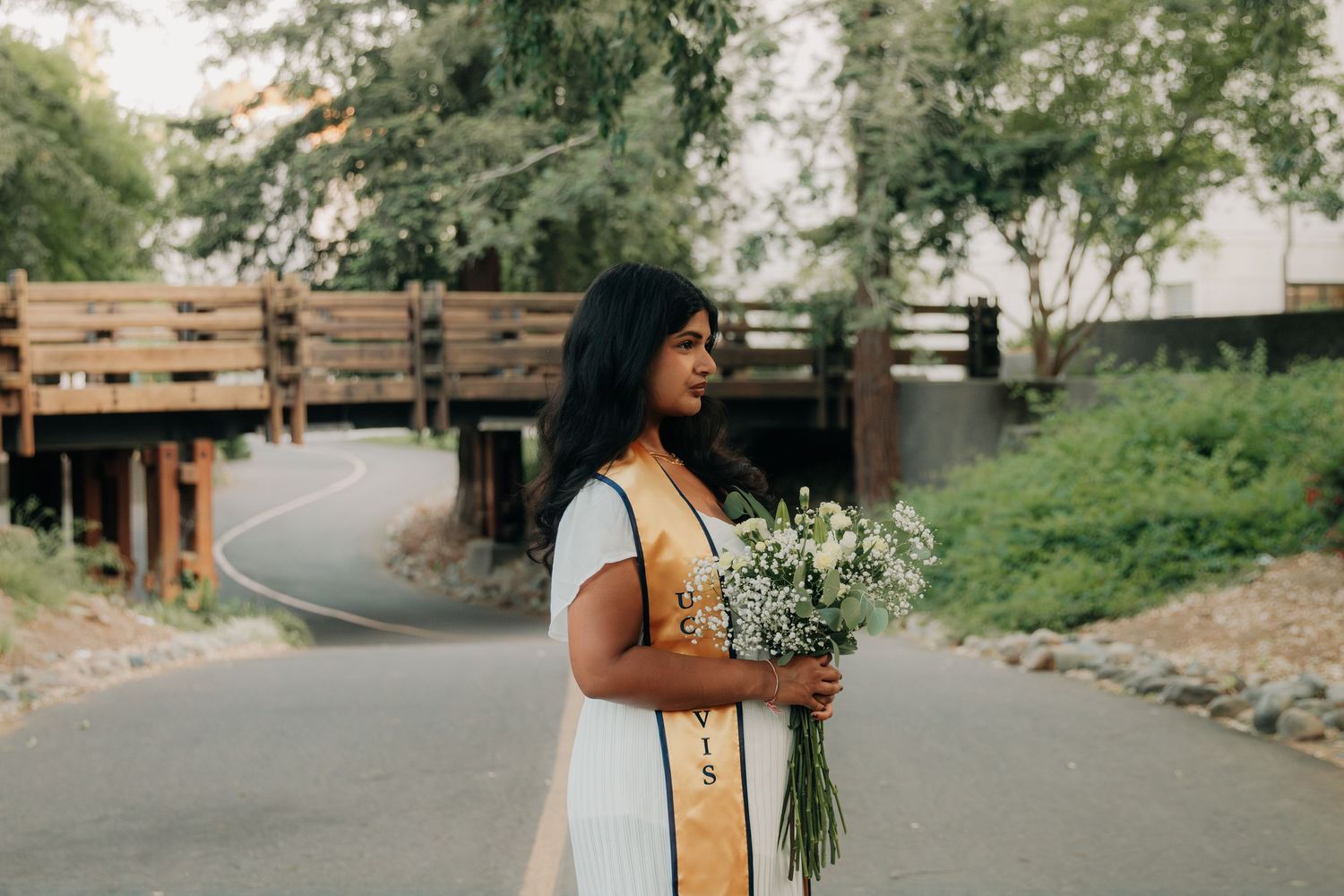 A person in a white dress stands on a winding path holding a bouquet of white wildflowers in a garden setting.