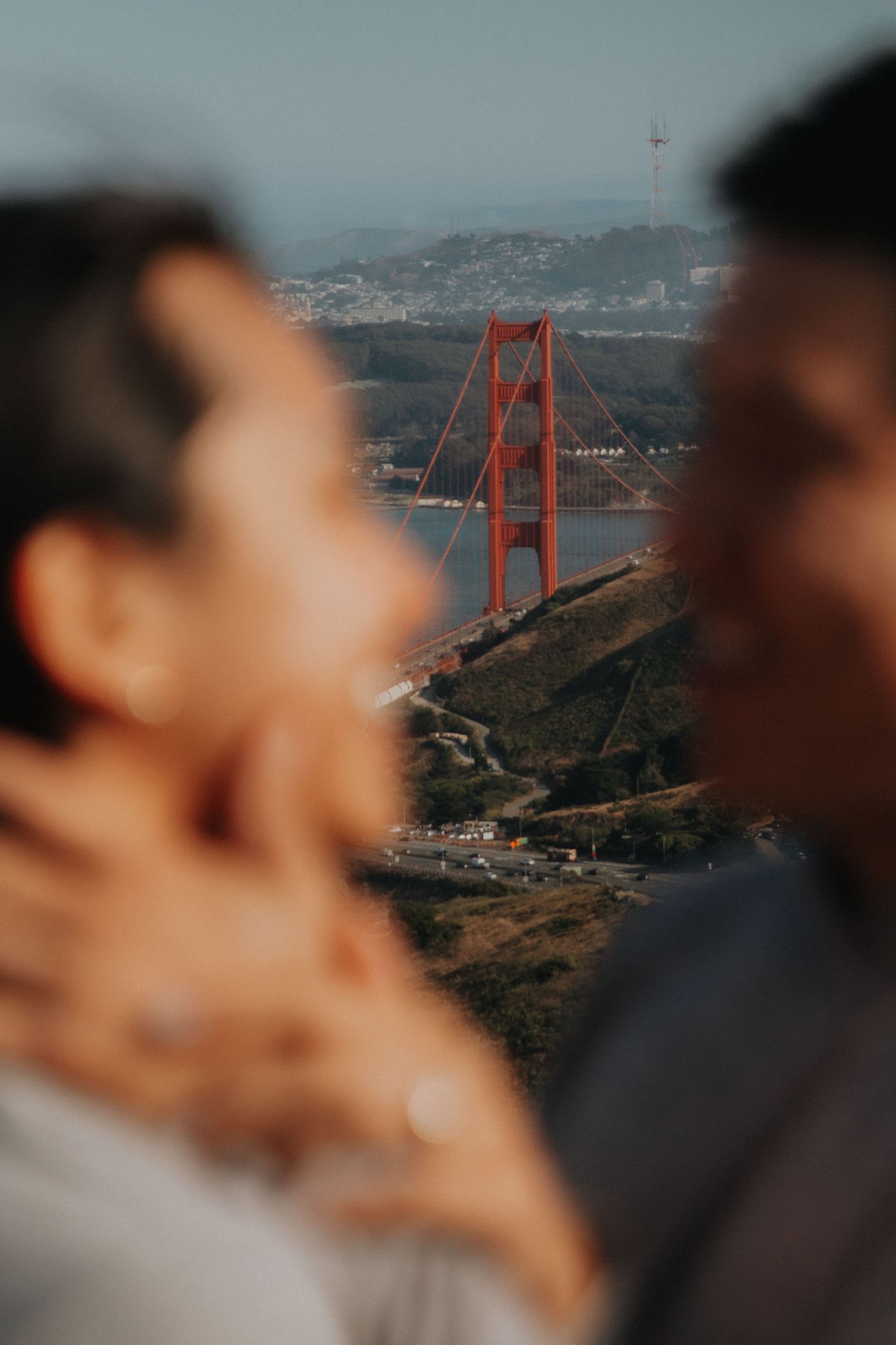 Blurred couple embraces in foreground with iconic Golden Gate Bridge and San Francisco Bay visible in background.