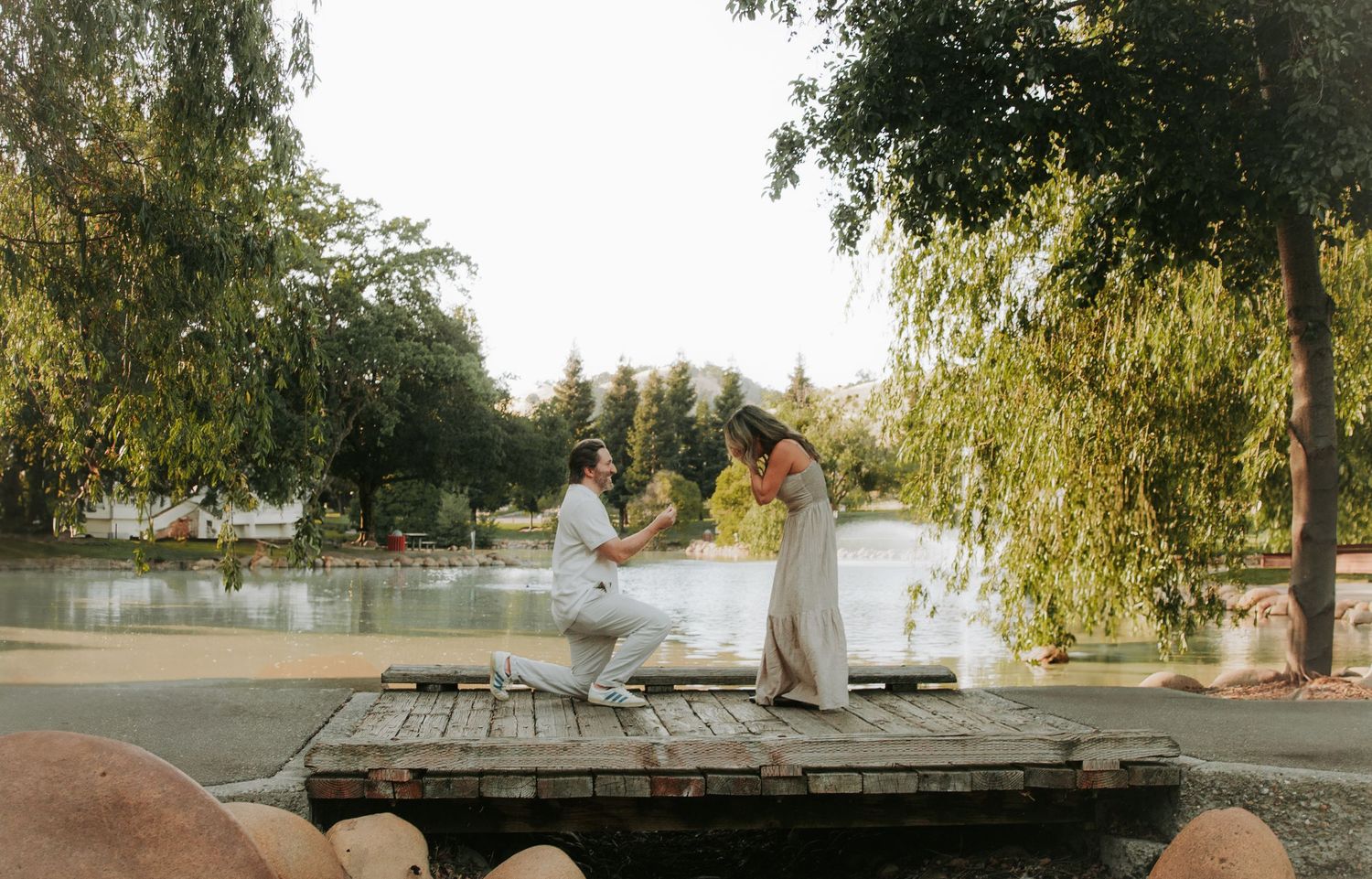 Two people in white dresses dance together on a wooden dock by a peaceful lake during golden hour.