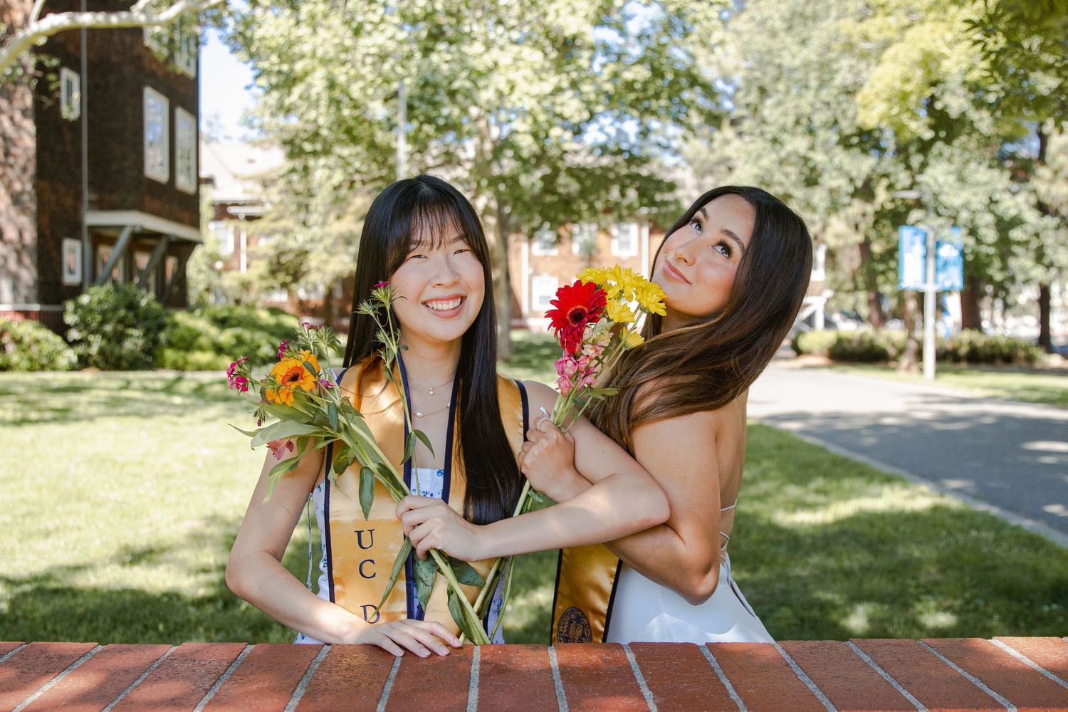 Two friends laughing while holding colorful flowers in a sunny garden.