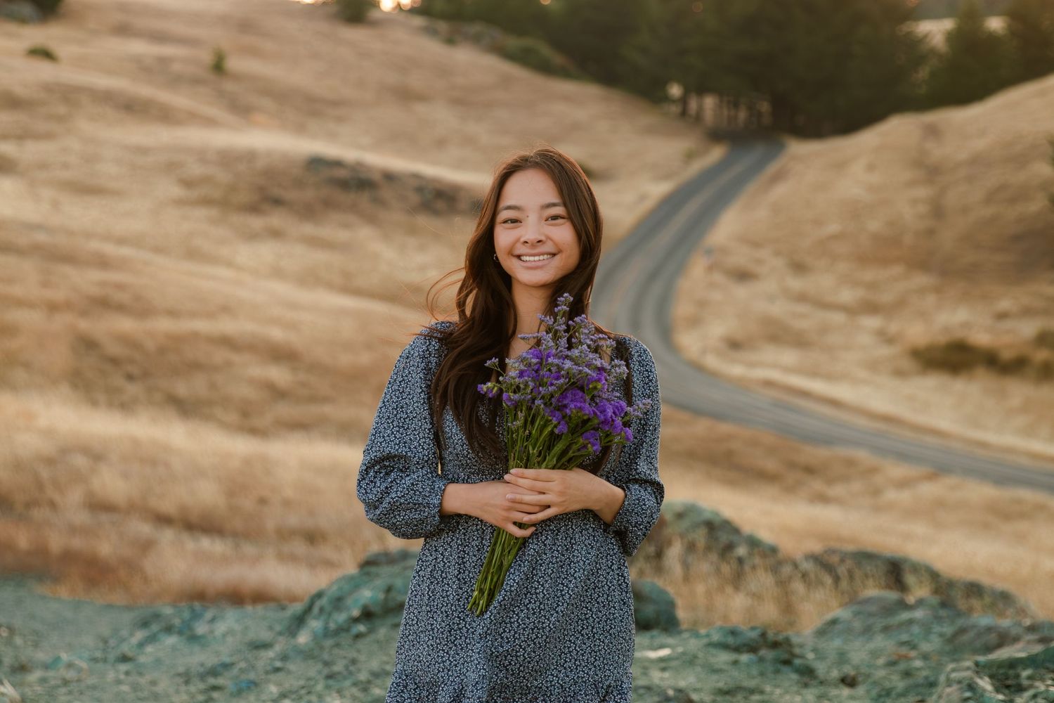 Person in blue denim jacket holding wildflowers on scenic mountain path.