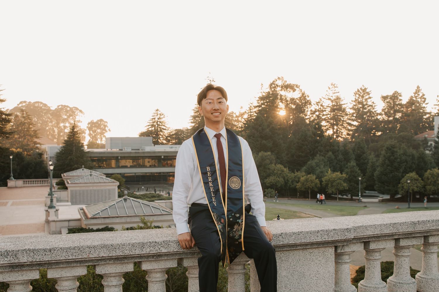 Graduate posing at sunset on a stone balcony surrounded by trees.
