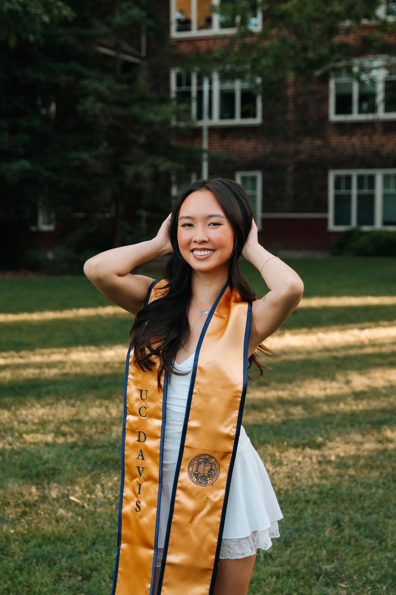 A graduate wearing white top and golden sash poses joyfully outdoors with brick building in background.
