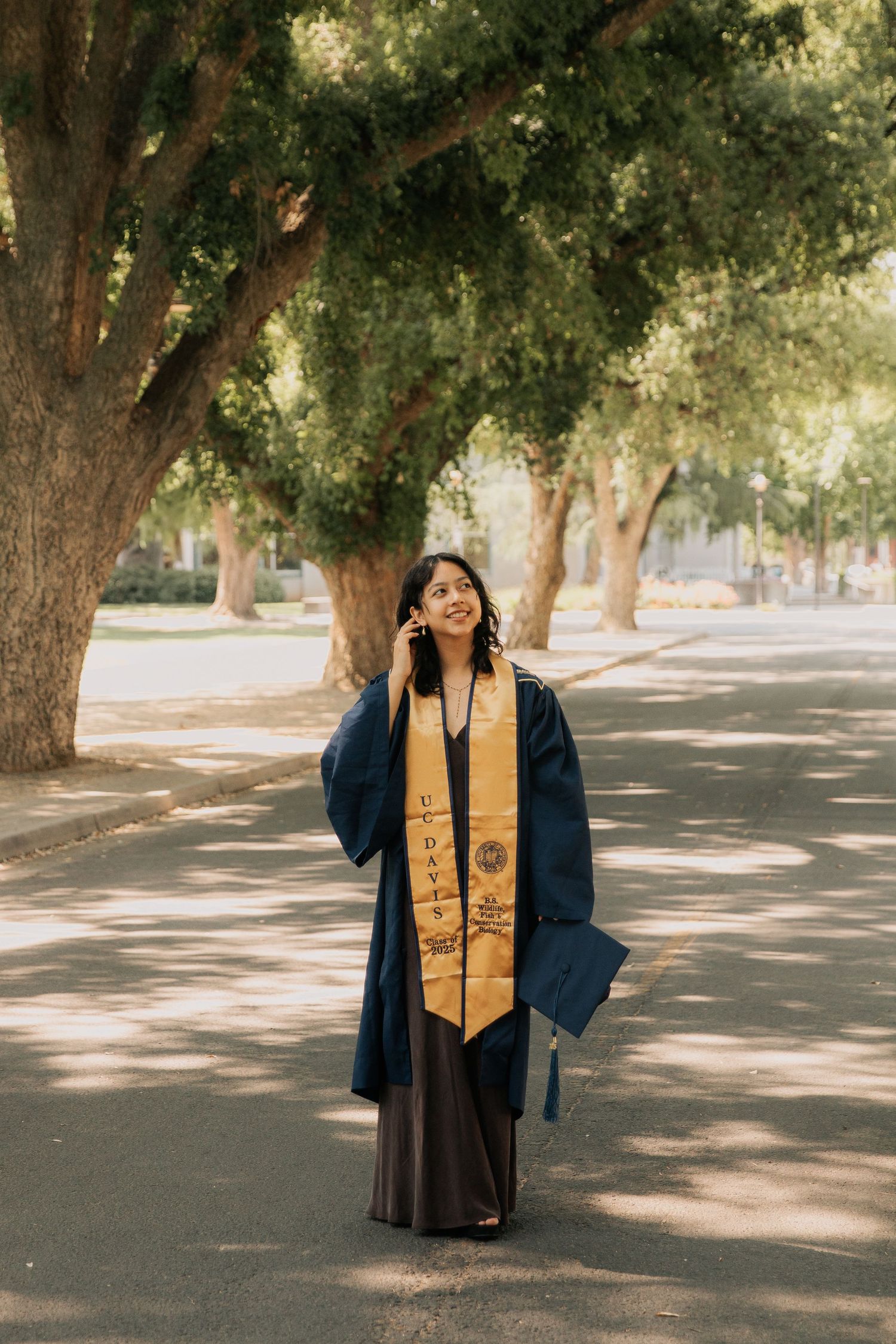 A graduate in dark gown and gold stole poses for photos under a shaded tree-lined pathway on graduation day.