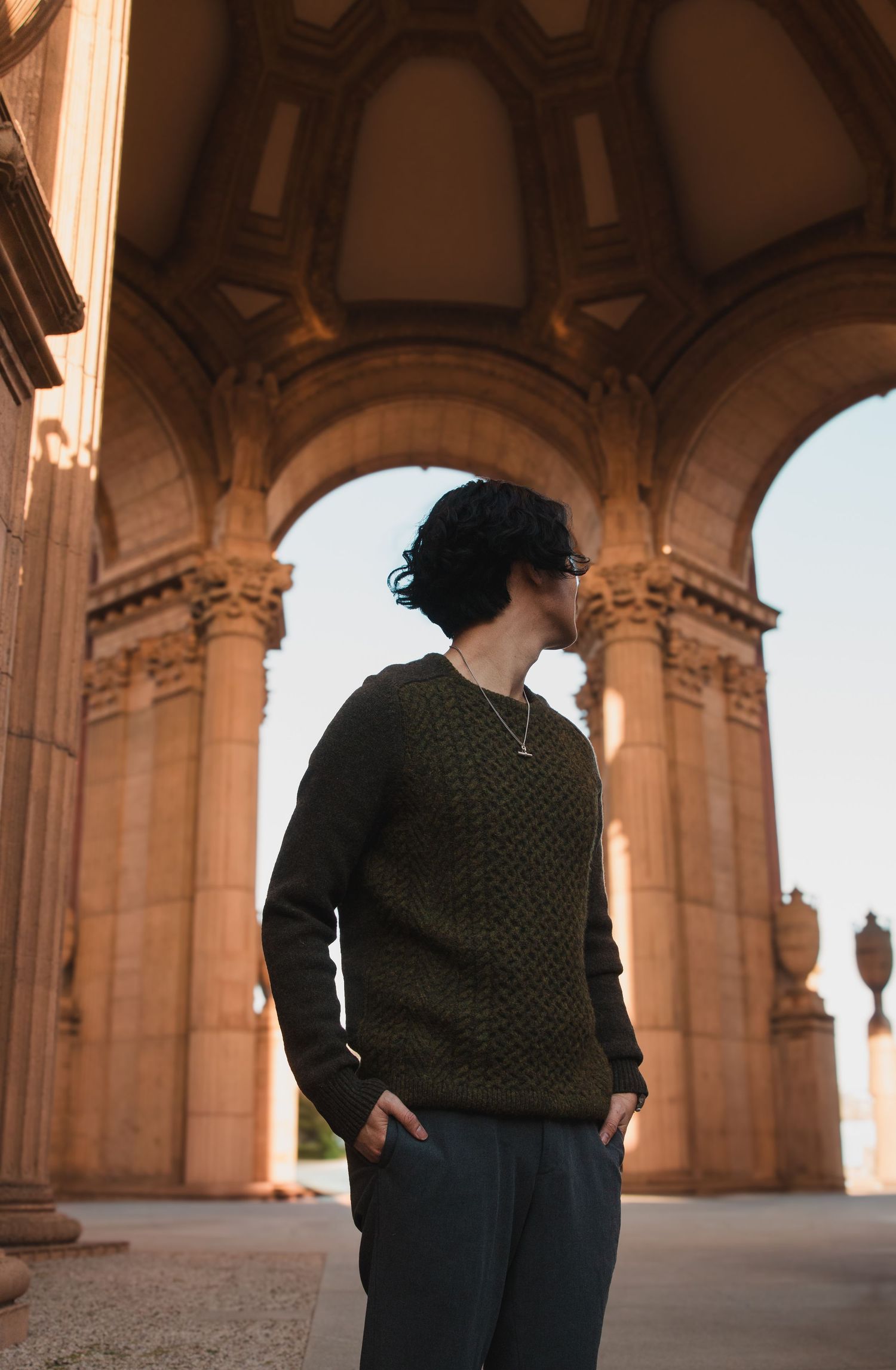 Person in dark sweater stands under ornate stone archway at sunset.