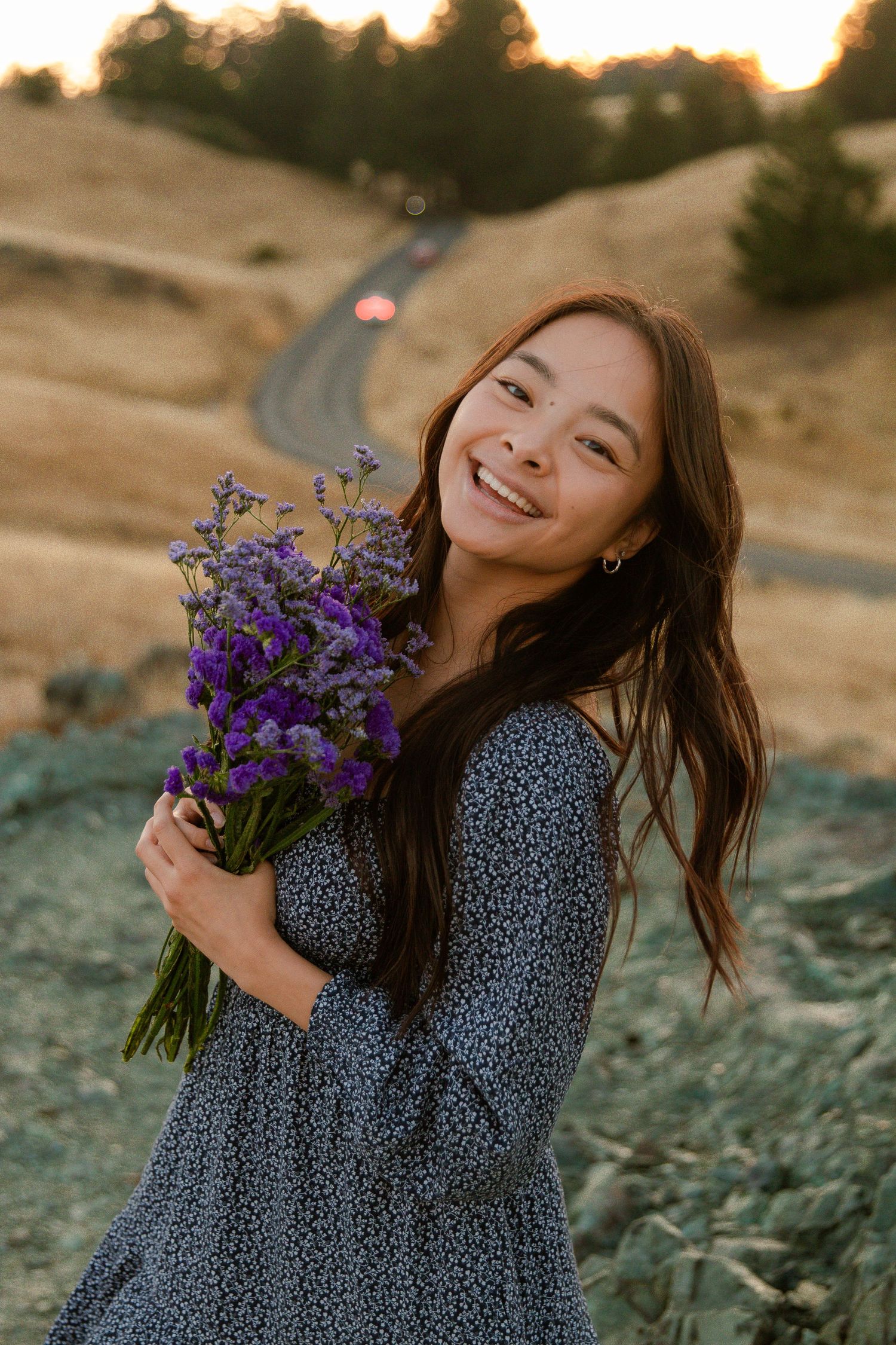 A woman in a floral dress holds purple wildflowers while smiling at sunset.