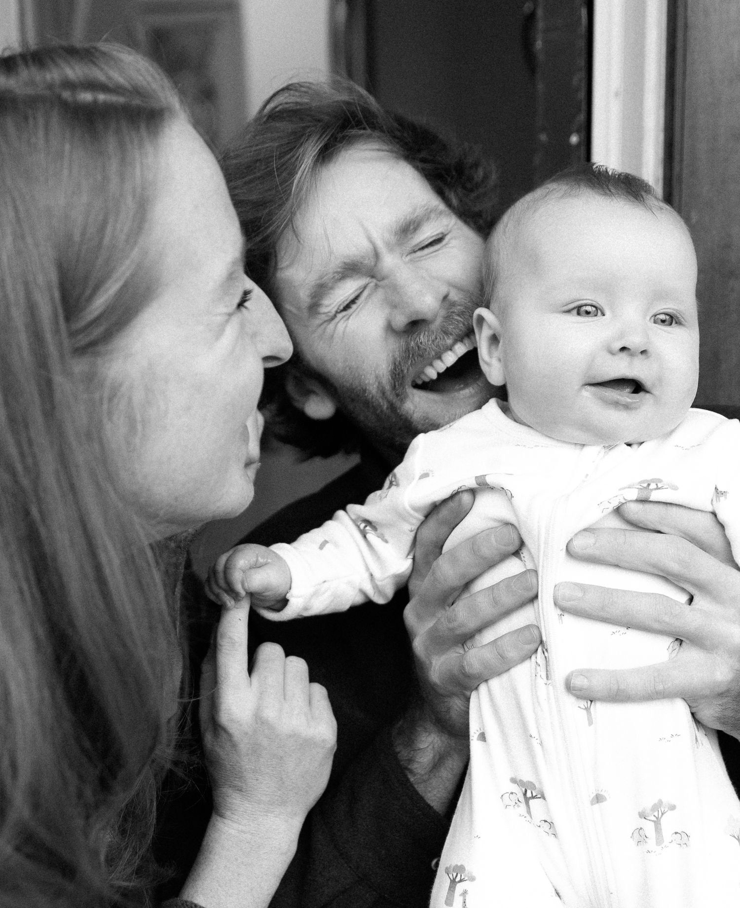 A candid black and white photograph captures a joyful family moment with a smiling baby in white clothing.