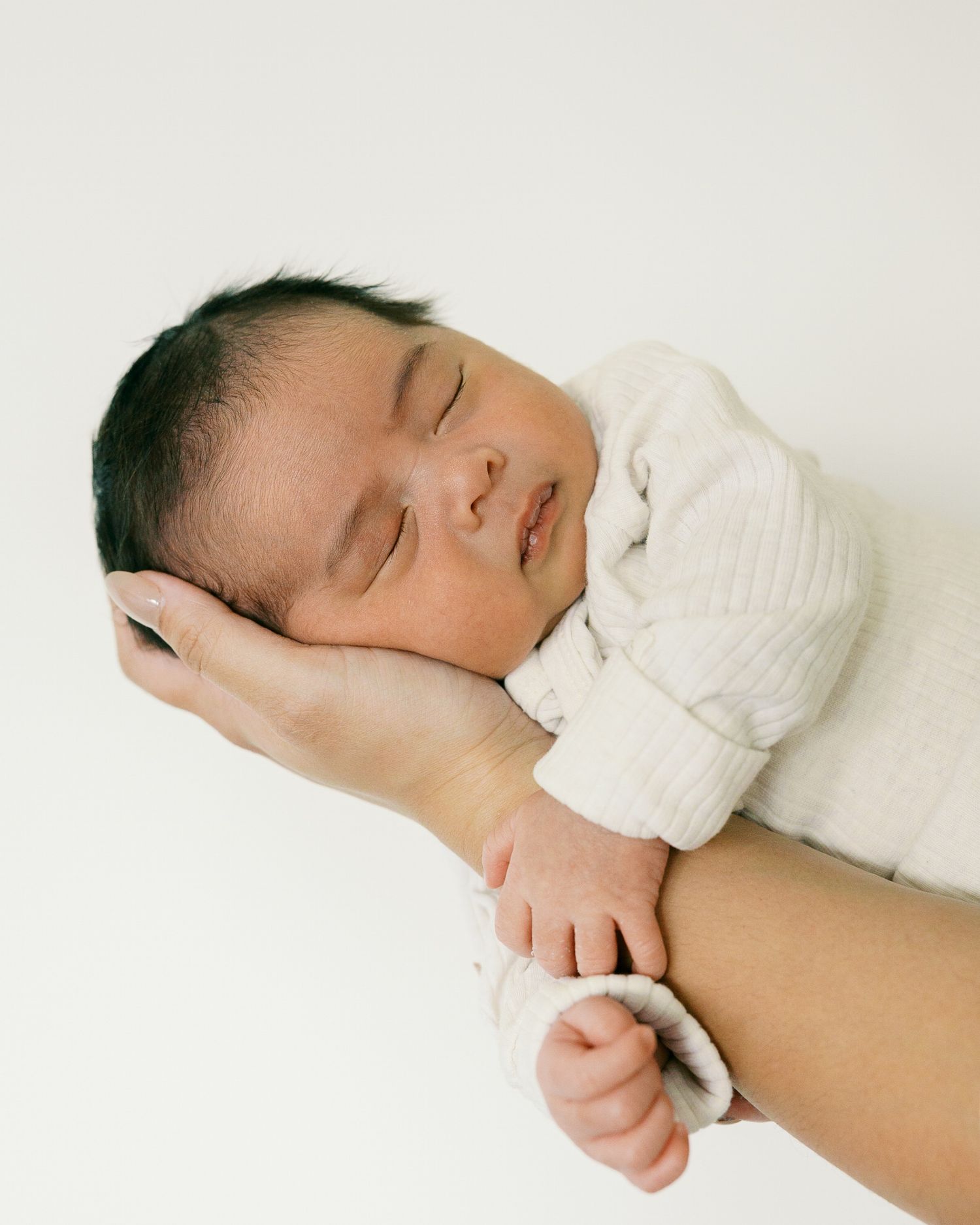 A peacefully sleeping newborn baby swaddled in white fabric rests gently on an adult's supportive hand.