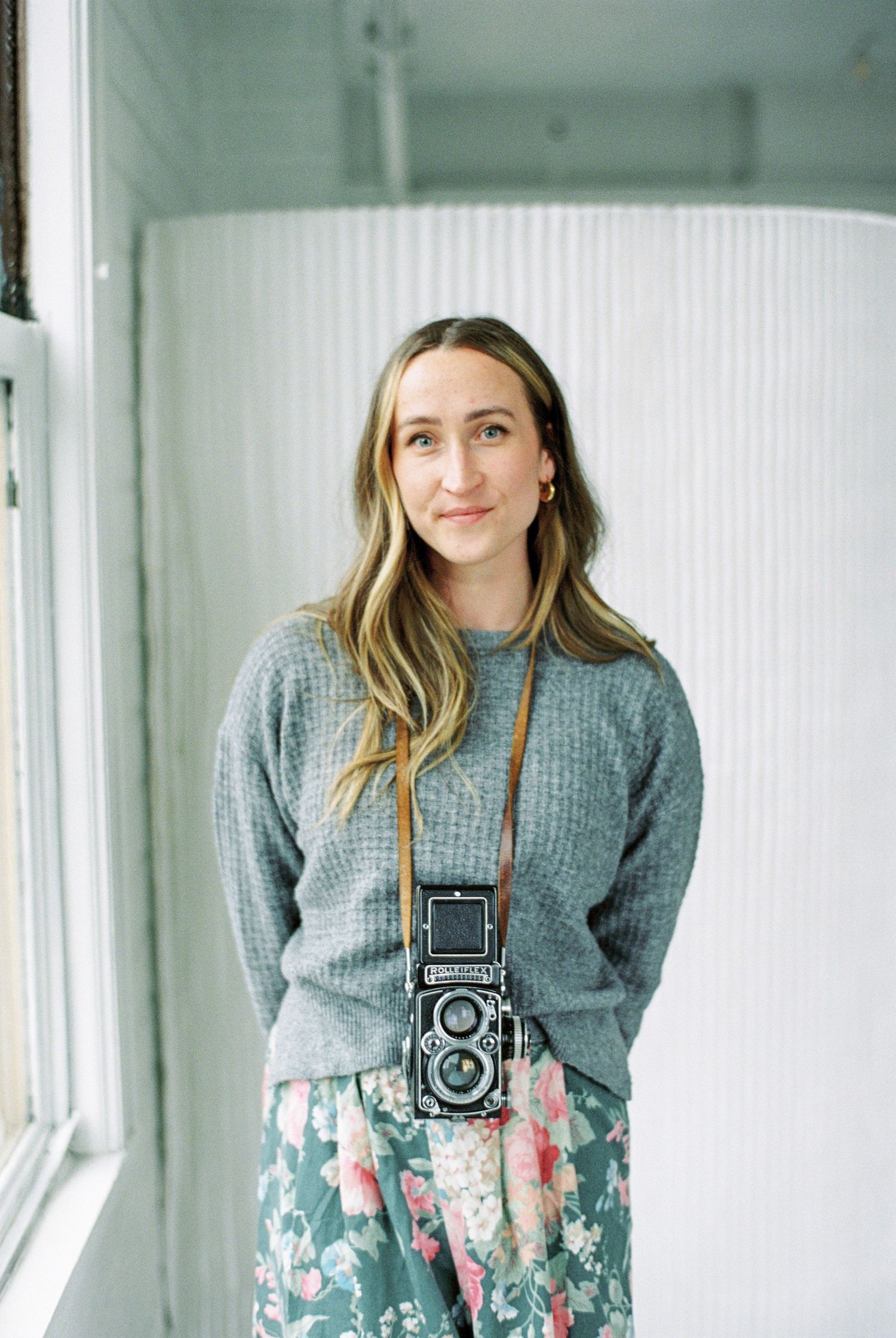 A photographer wearing a grey knit sweater and floral skirt poses with a vintage Rolleiflex camera around their neck.