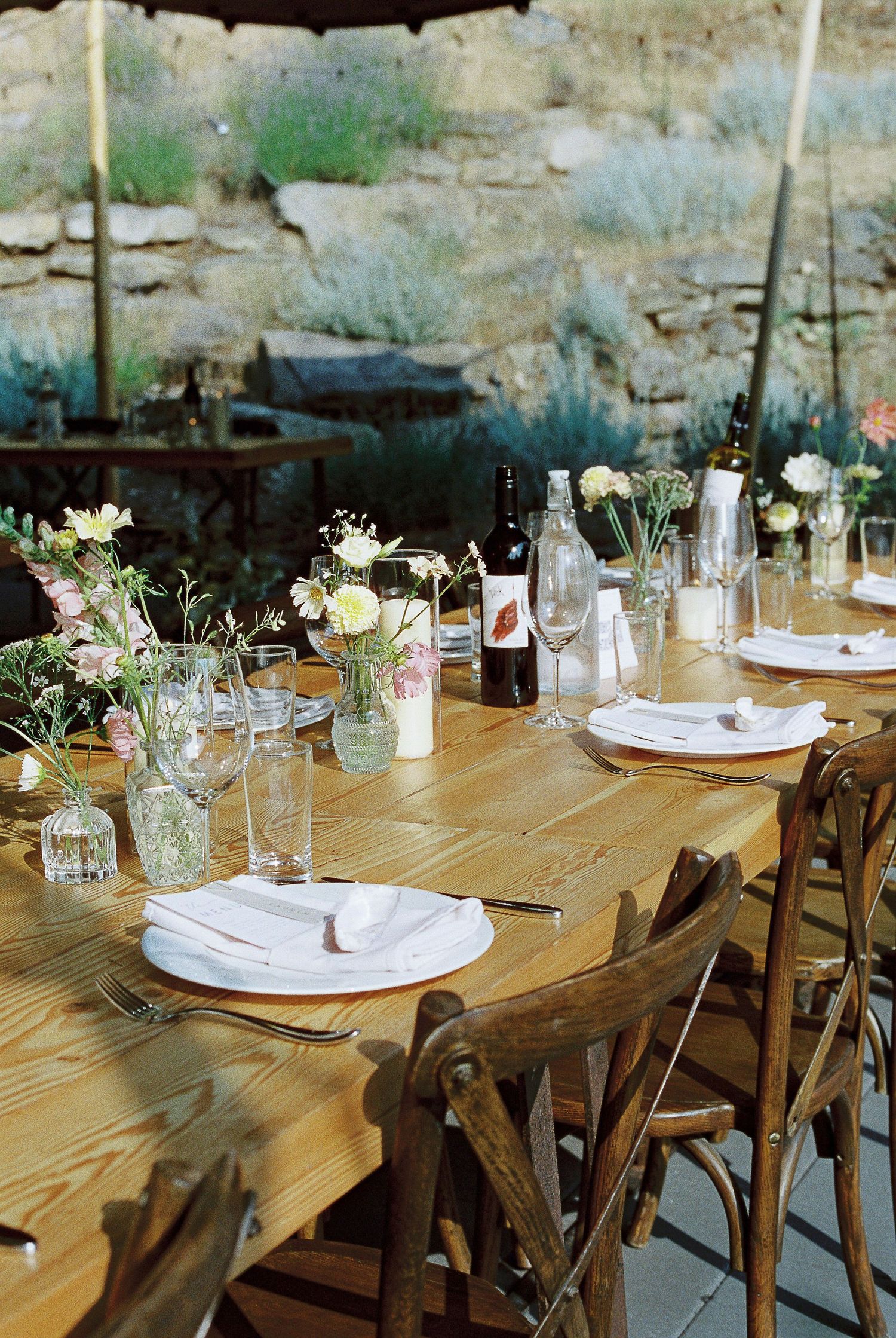 A rustic wooden dining table set for an outdoor event with wildflower centerpieces, wine bottles and vintage wooden chairs.