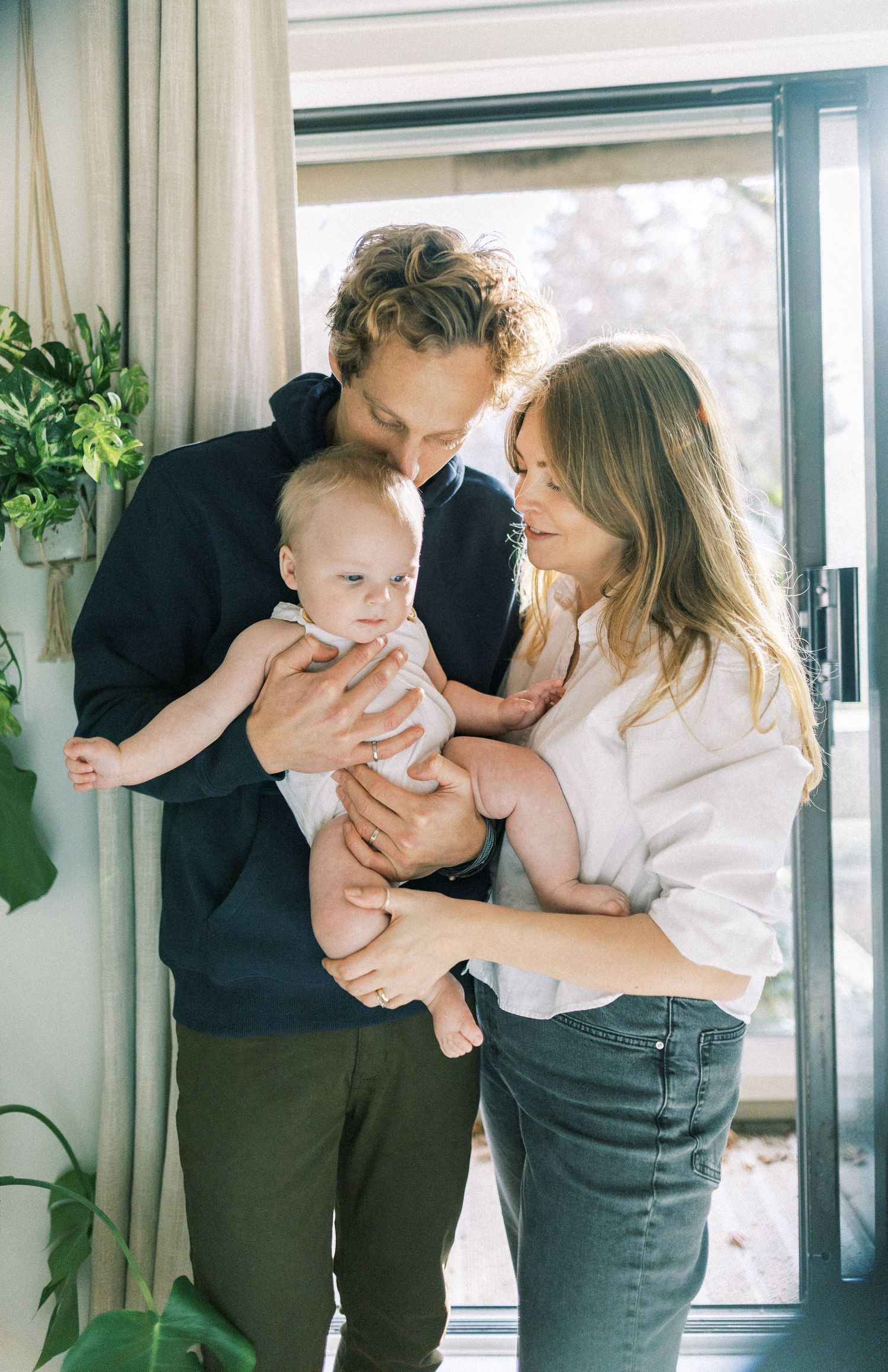 Family stands together in natural light by patio door with small infant in casual indoor setting.