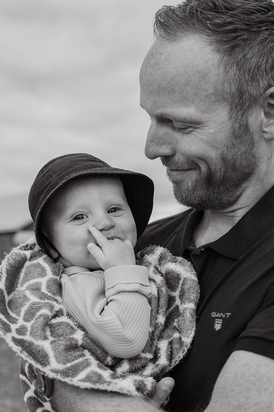 Svartvit bild på leende pappa hållandes litet barn i famnen med handen i munnen insvept i en filt och bärande solhatt. Familjefotograf, Umeå