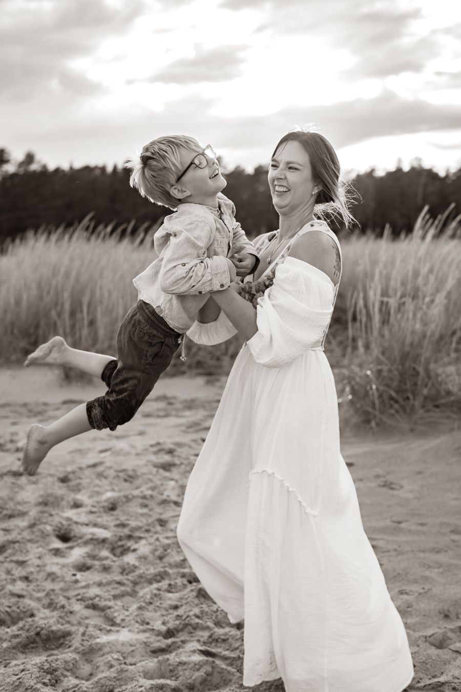 Svartvit bild med en glädje och lek mellan mamma och son på sandstranden med högt gräs i bakgrunden. Fotograf, Familjefotograf, Umeå
