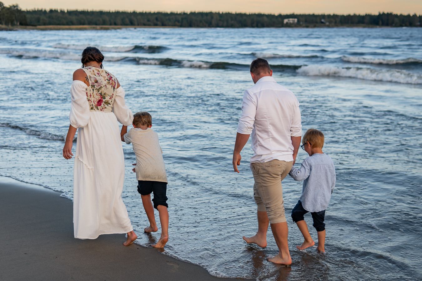 En familj i ljusa kläder går barfota hand i hand vid strandkanten i solnedgången när vågorna rullar in. Familjefotograf, Umeå, Västerbotten