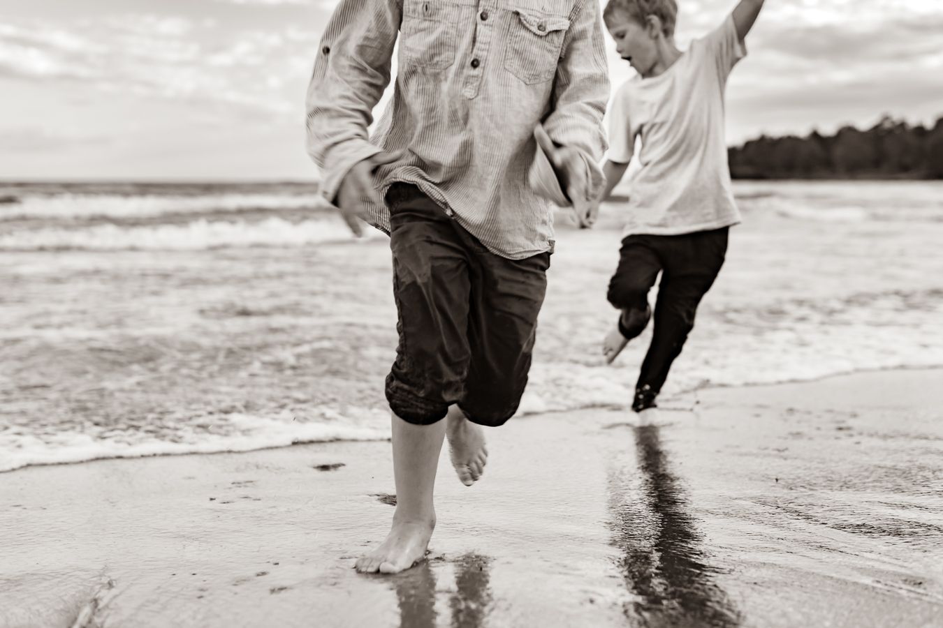 Svartvit bild på två barn som springer och leker i vattenbrynet på en sandstrand med havet i bakgrunden. Fotograf, Familjefotograf, Umeå