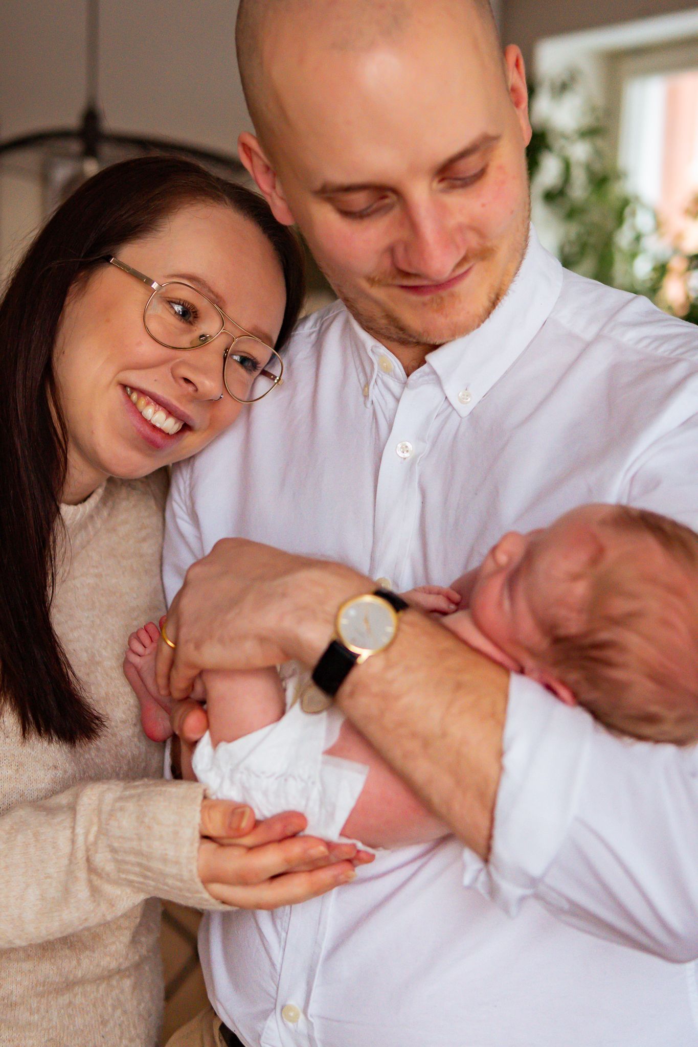 Föräldrar ler och håller varsamt och kärleksfullt sin nyfödda bebis i famnen i ett varmt naturligt ljus inomhus. Nyföddfotograf, Umeå