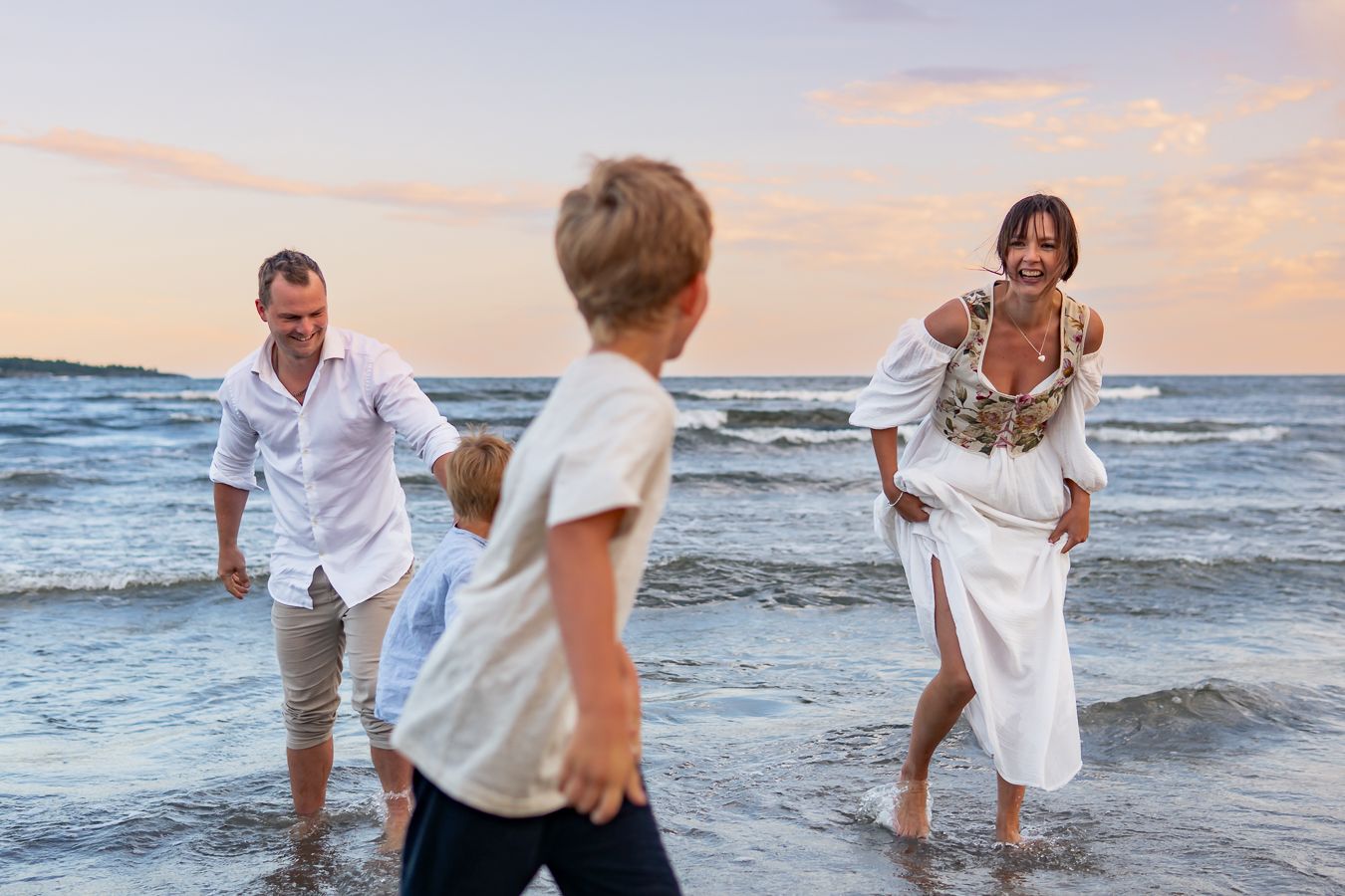 En familj leker tillsammans i vattnet vid strandkanten i solnedgång med havet i bakgrunden. Fotograf, Familjefotograf, Umeå