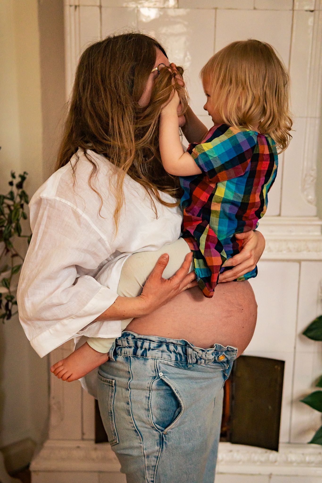 En gravid kvinna i vit skjorta och jeans håller en liten dotter i famnen med färgglad klänning bredvid en kakelugn. Gravidfotograf, Umeå