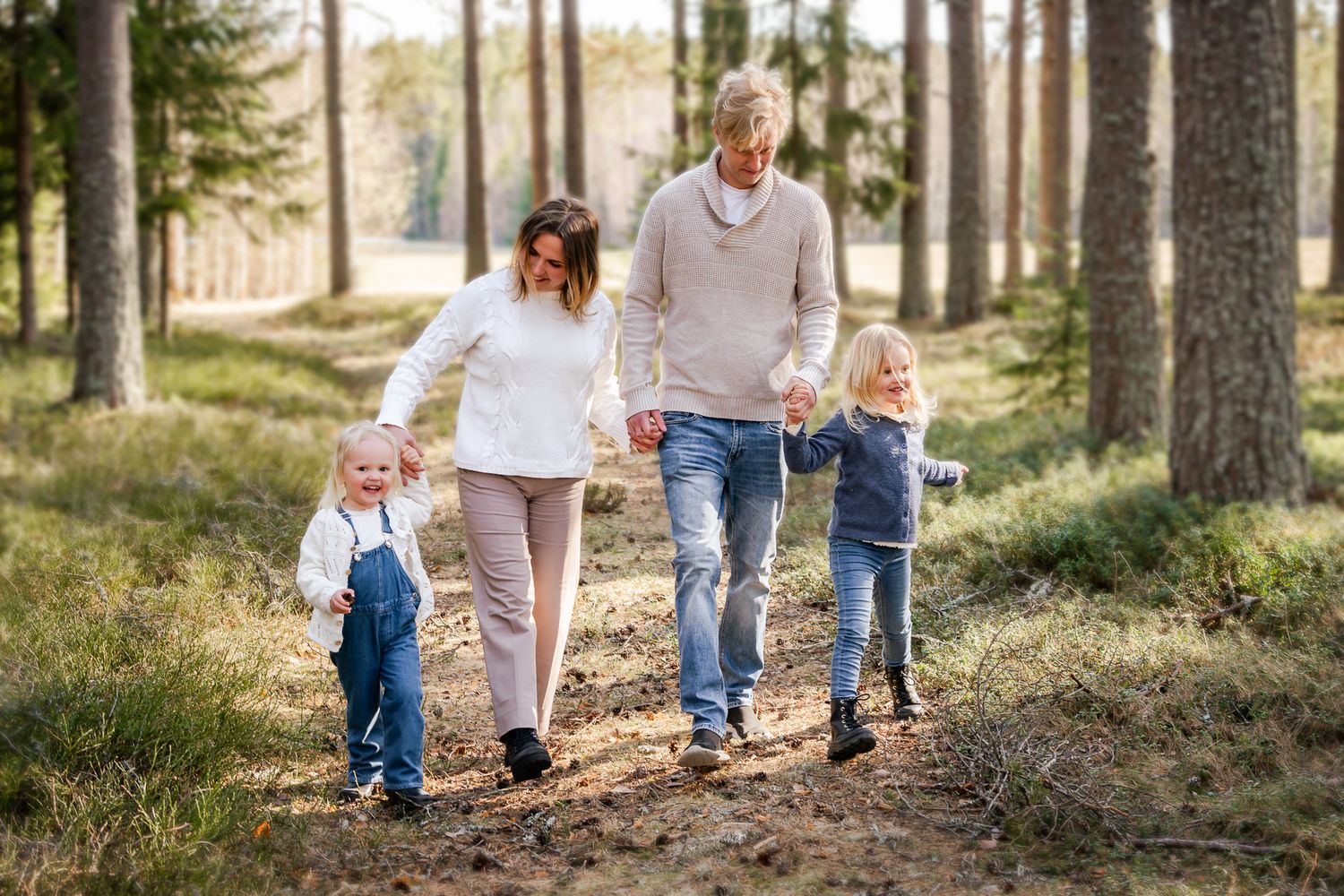 Familj med två barn går hand i hand på en bred stig i skogen där solen lyser in mellan träden. Fotograf, famijefotograf, Umeå, Västerbotten