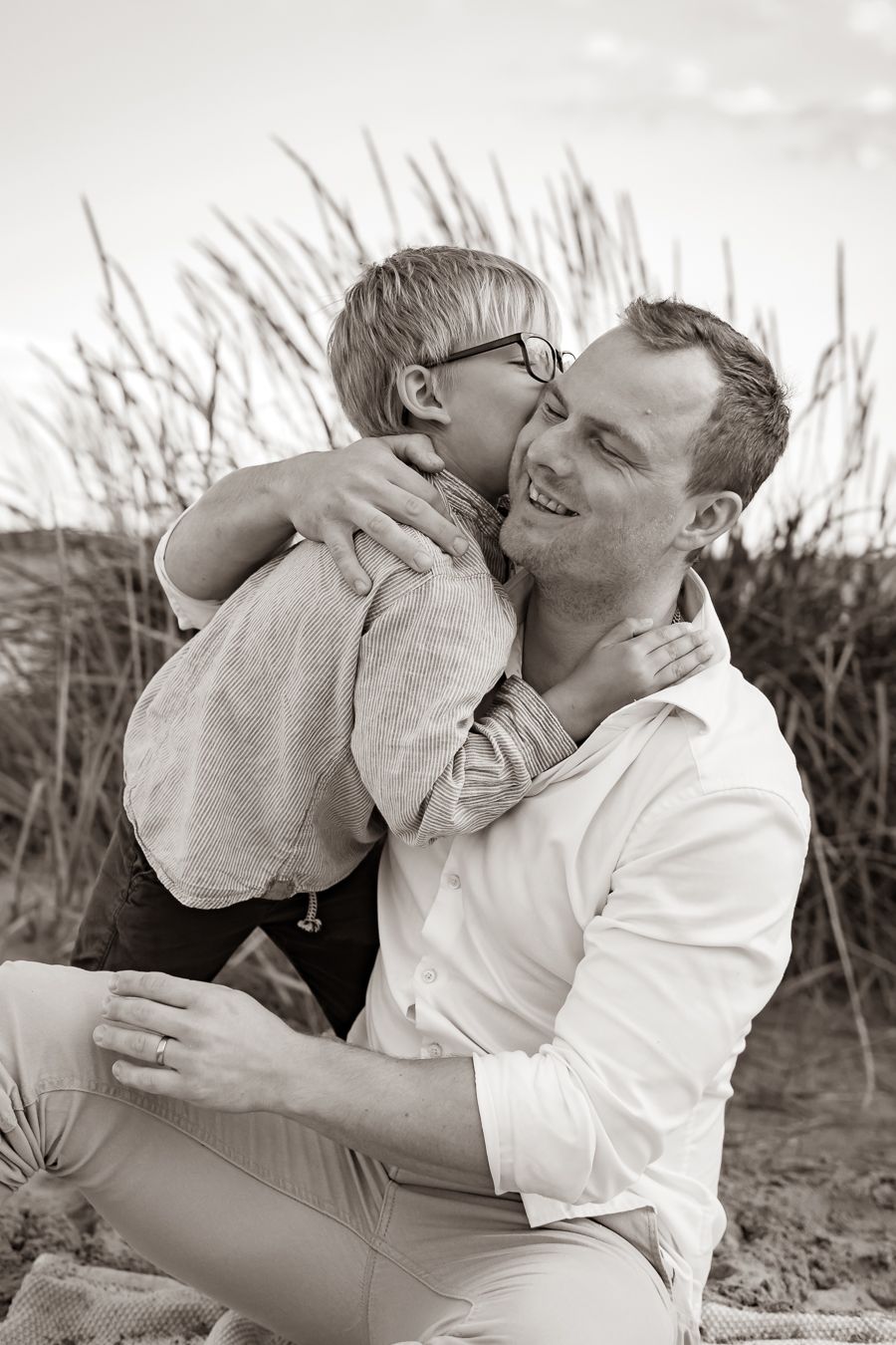 Kärleksfull svartvit bild av en pappa och son som kramas på stranden med högt gräs i bakgrunden. Familjefotograf, Umeå, Västerbotten