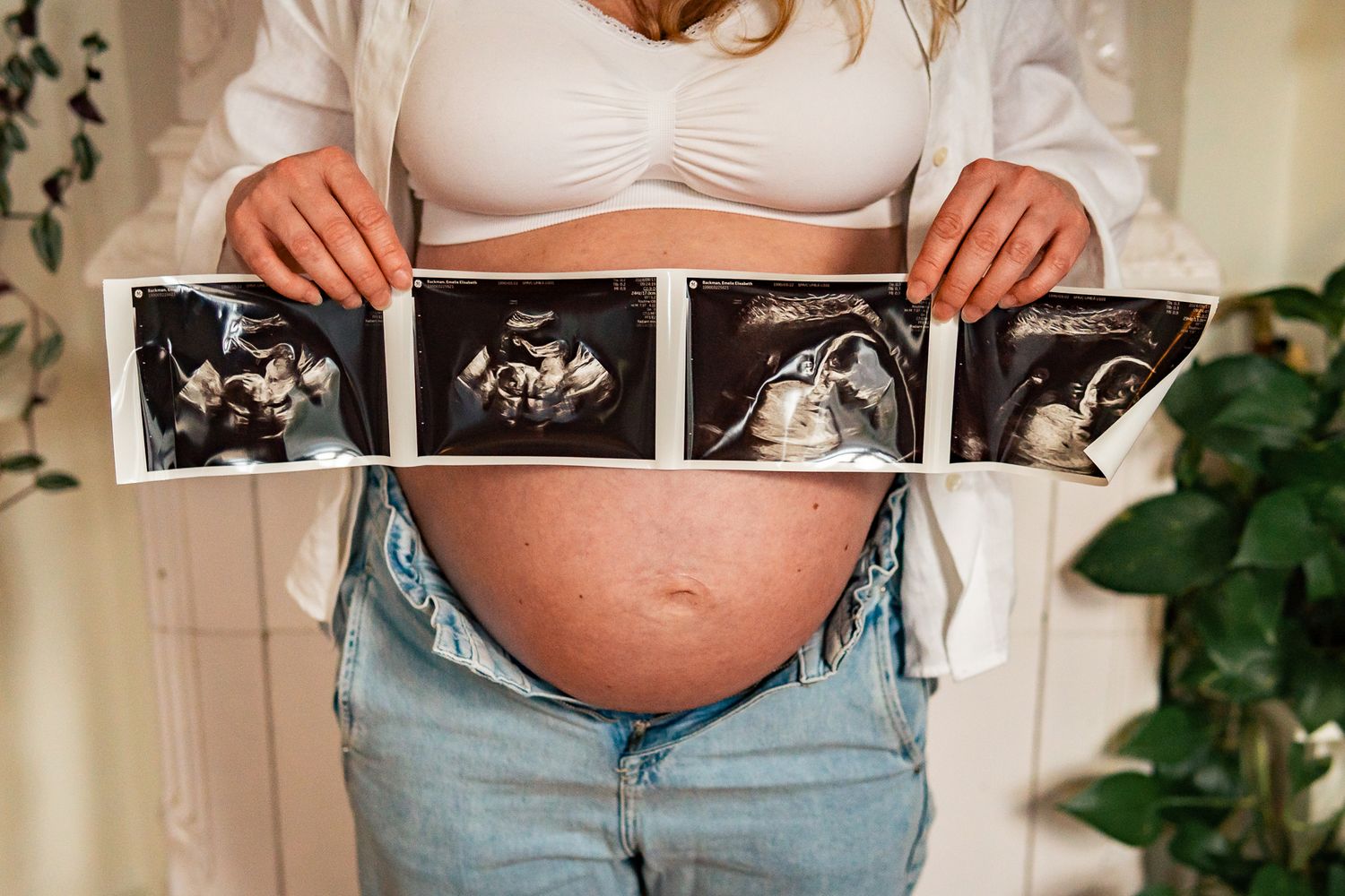 Gravid kvinna i vit bh och jeans håller ultraljudsbilder framför gravida magen. Gravidfotograf, Umeå, Västerbotten