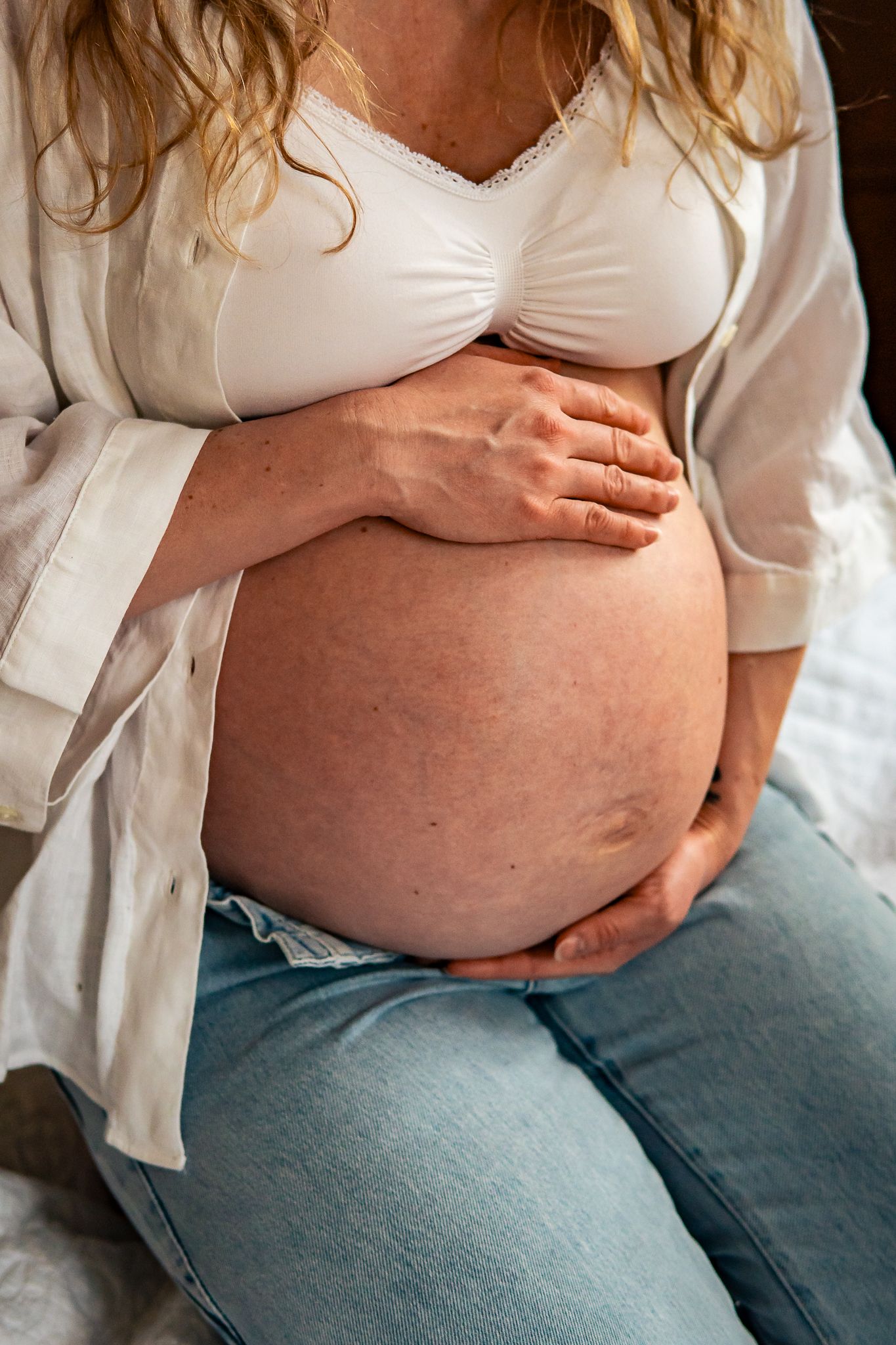 Gravid kvinna i vit bh och jeans håller sin gravida mage mellan sina händer. Gravidfotograf, Umeå, Västerbotten, Fotograf