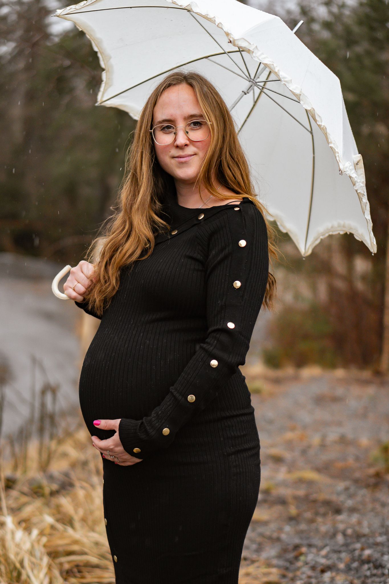 Gravid kvinna i svart klänning med guldknappar står i regnet med ett vitt spetsparaply och håller om sin gravida mage. Gravidfotograf, Umeå
