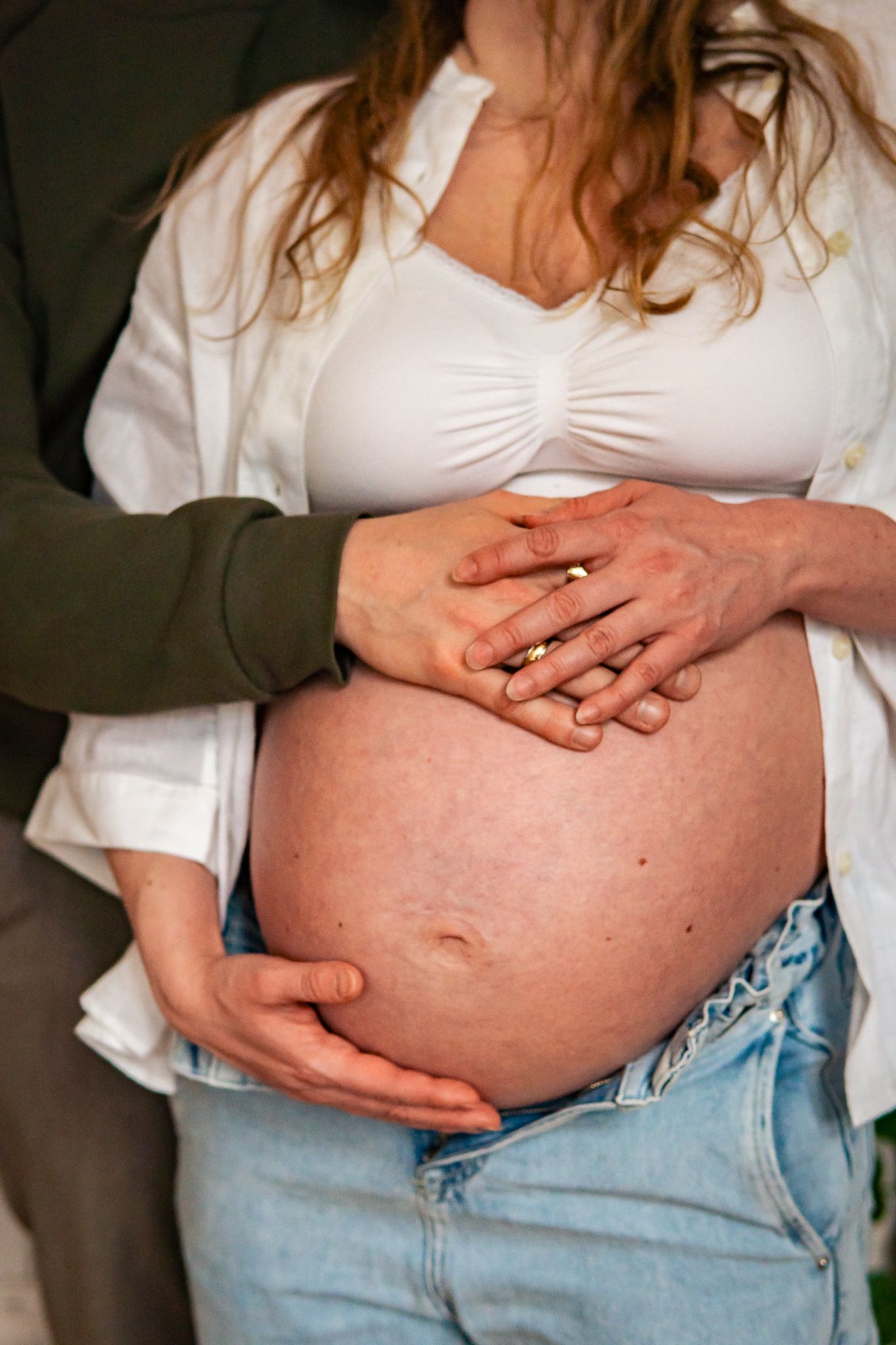 Närbild på händer som vilar på en gravid mage, kvinnan bär jeans och en vit skjorta. Gravidfotograf, Umeå, Västerbotten