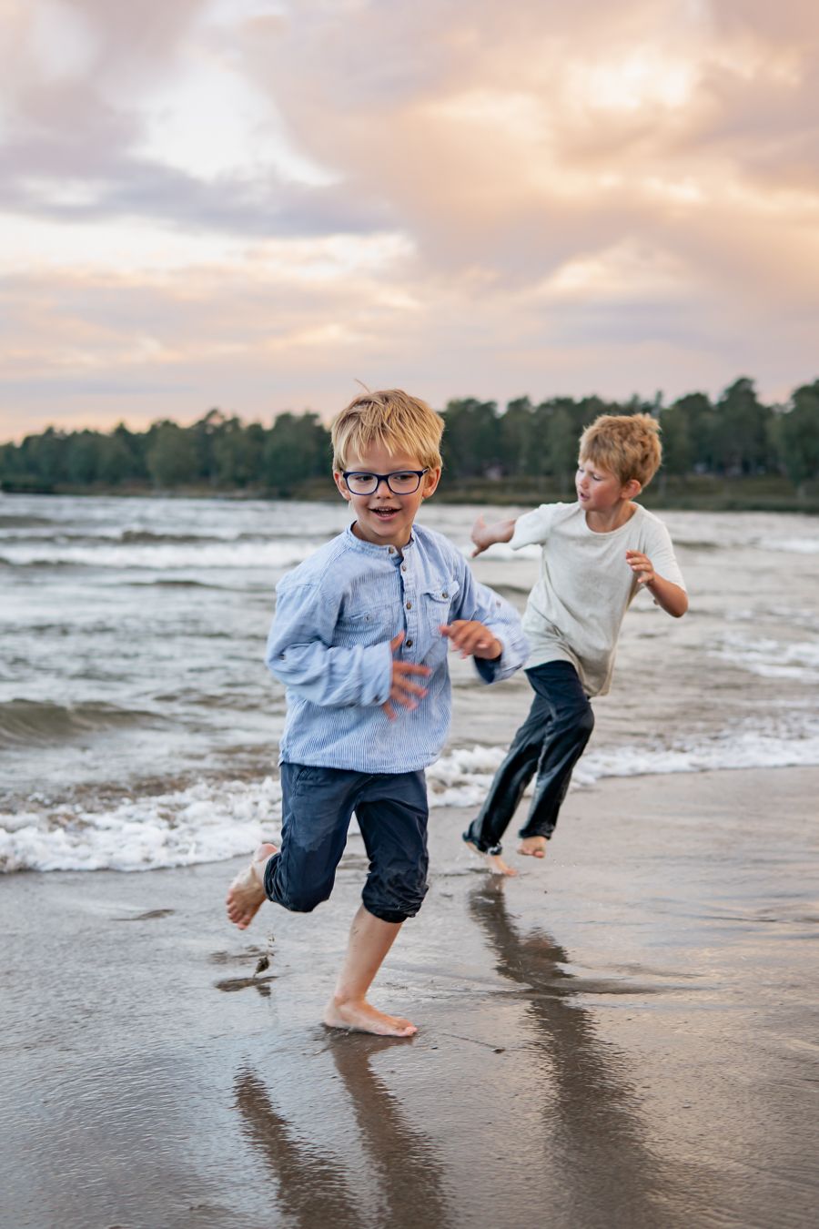Två barn springer barfota längs vattenbrynet på sandstranden i solnedgången med havet i bakgrunden. Familjefotograf, Umeå, Västerbotten