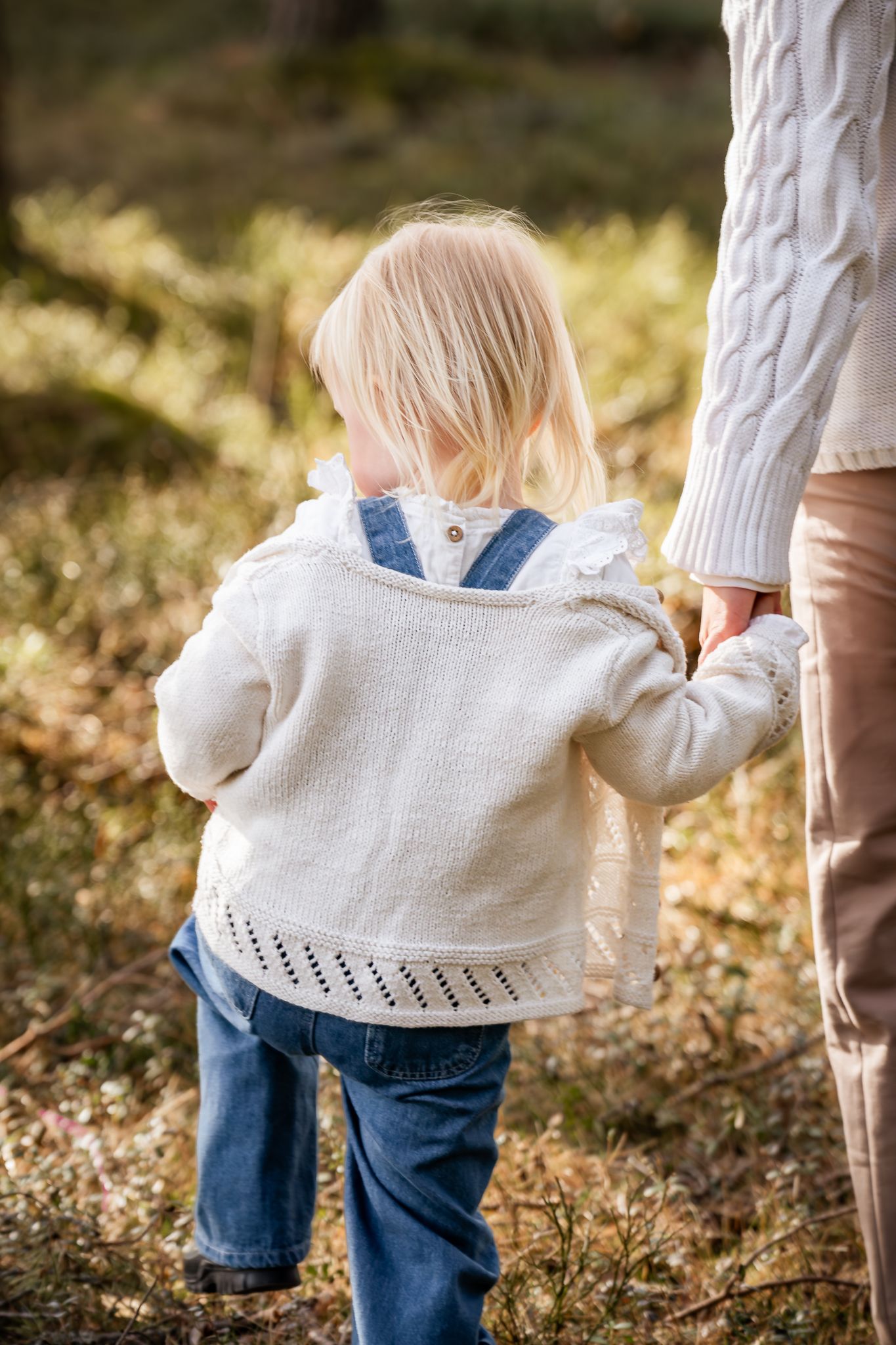 Bild bakifrån på liten tjej som håller mammas hand när hon går genom snårig skog och solen lyser varmt. Fotograf, familjefotograf, Umeå