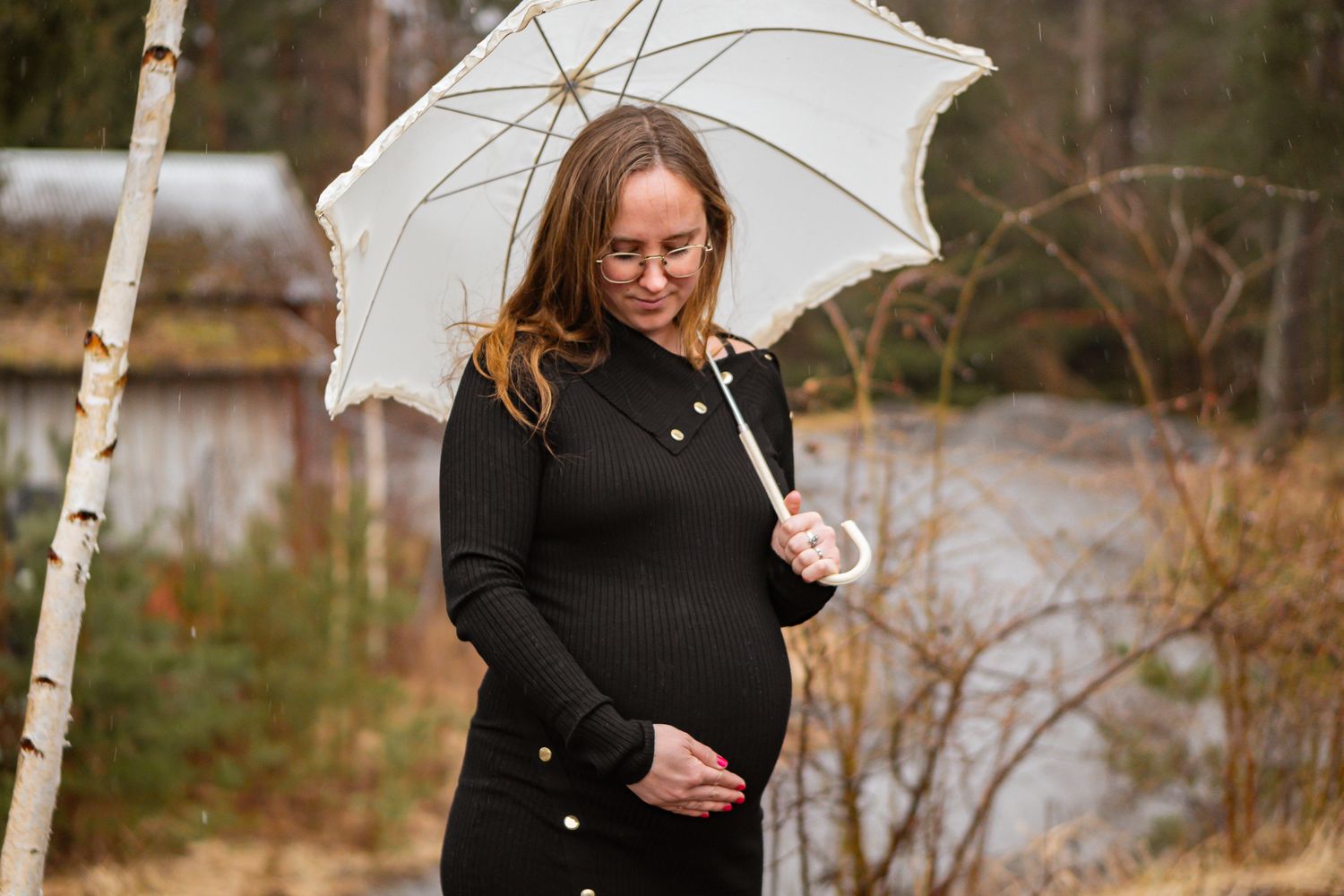 Gravid kvinna i svart klänning med guldknappar står ute med vitt spetsparaply, tittar ner på gravida magen hon håller. Gravidfotograf, Umeå