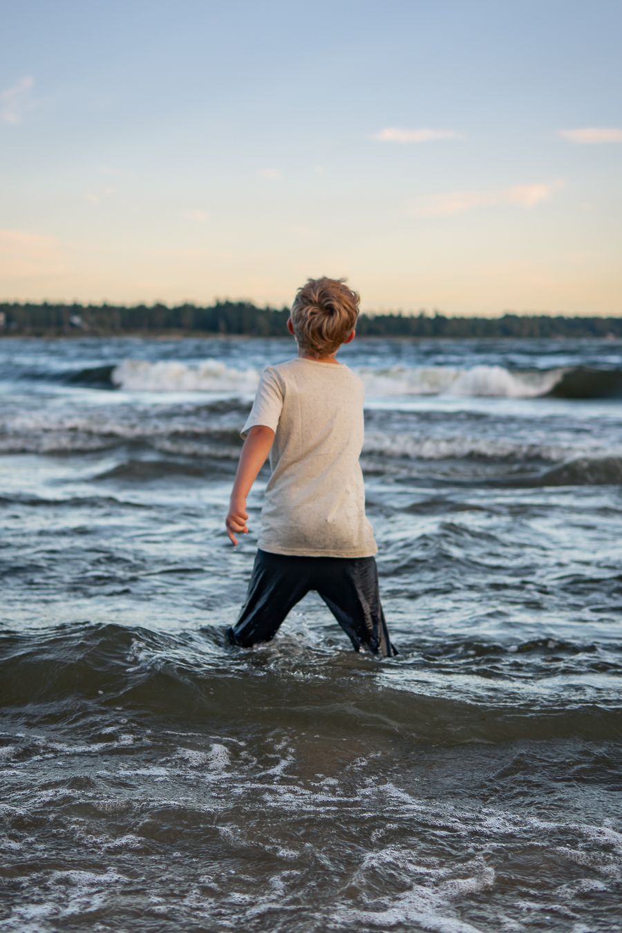 En ung pojke står med kläderna på i havets vågor vid strandkanten i solnedgången. Familjefotograf, Umeå, Västerbotten