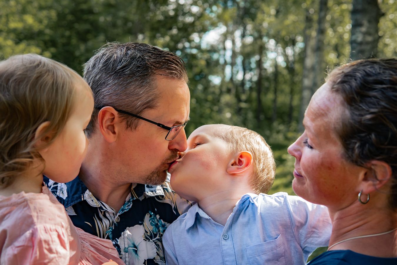 En familj delar närhet och pussar utomhus en solig sommardag med träd i bakgrunden. Fotograf, Familjefotograf, Umeå, Västerbotten