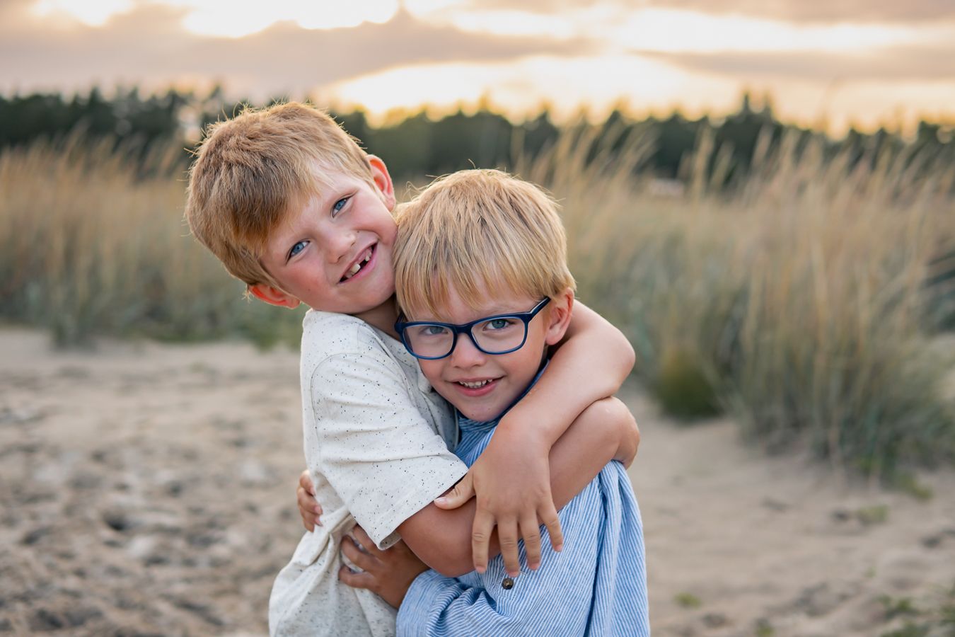 Två syskon delar en kram med glädje och kärlek på en sandstrand i solnedgång med högt gräs i bakgrunden. Fotograf, Familjefotograf, Umeå