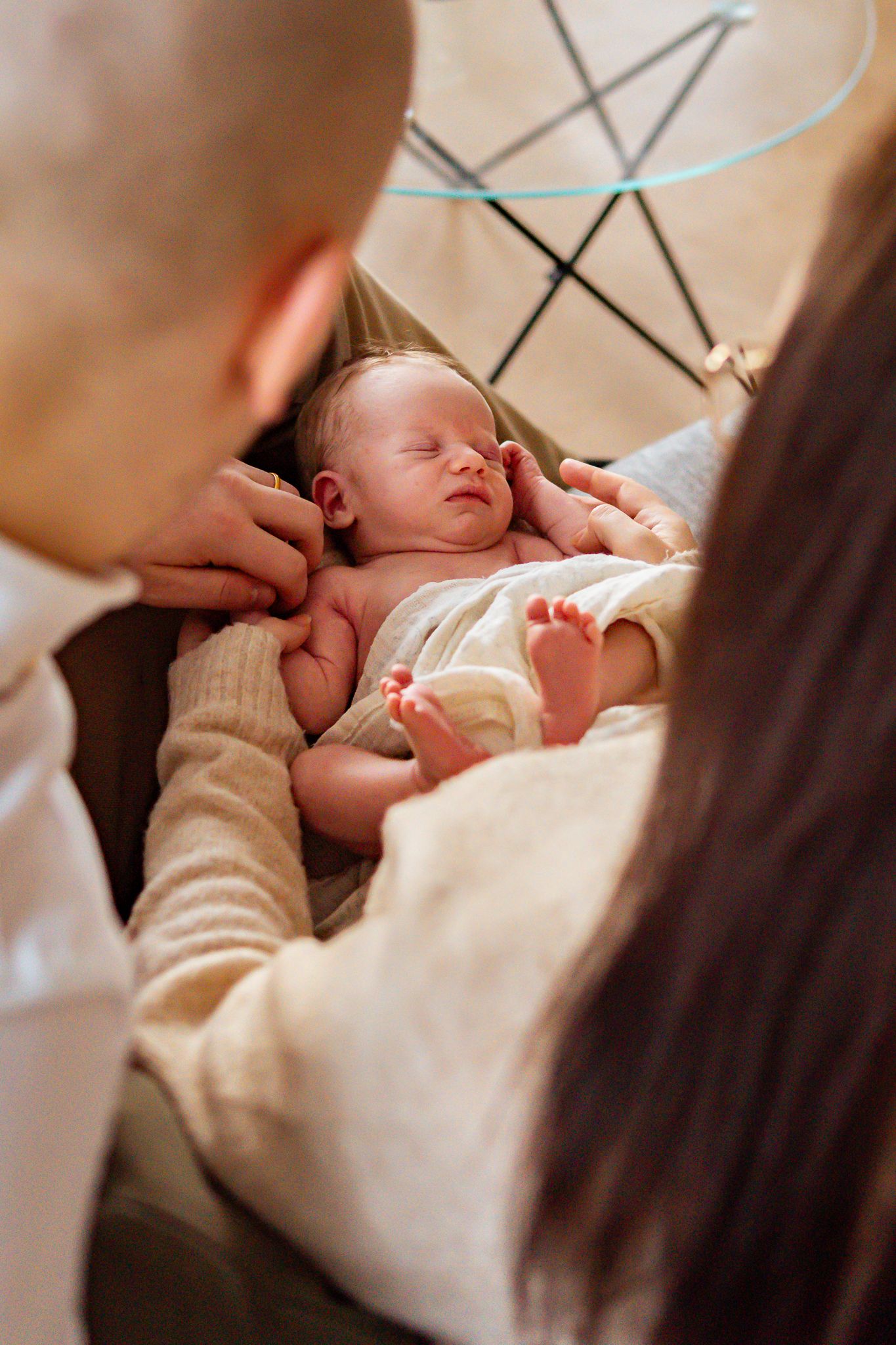 Nyfödd bebis sover fridfullt i en vit filt i mamma knä samtidigt som föräldrarnas händer varsamt håller i honom. Nyföddfotograf, Umeå