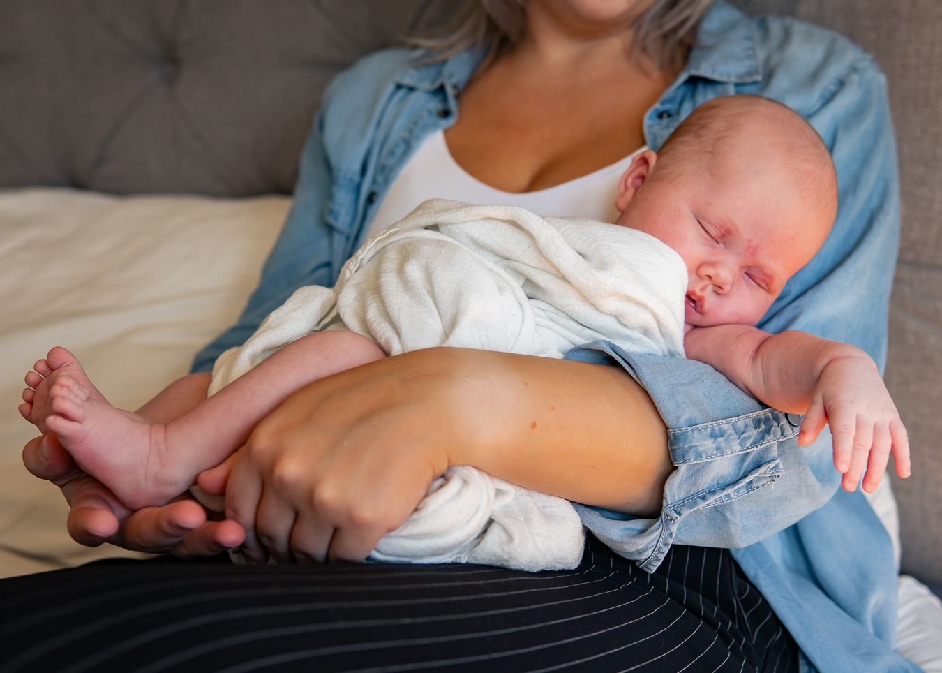 Sovande bebis i vit filt ligger i mammas famn som bär denim i mjukt naturligt ljus. Fotograf, nyföddfotograf, Umeå, Västerbotten