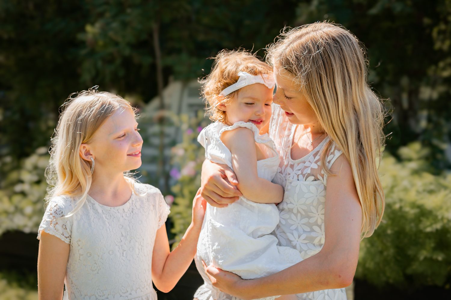Tre tjejer i vita spetsklänningar står tillsammans i trädgården en solig sommardag. Familjefotograf, Umeå, Fotograf, Västerbotten