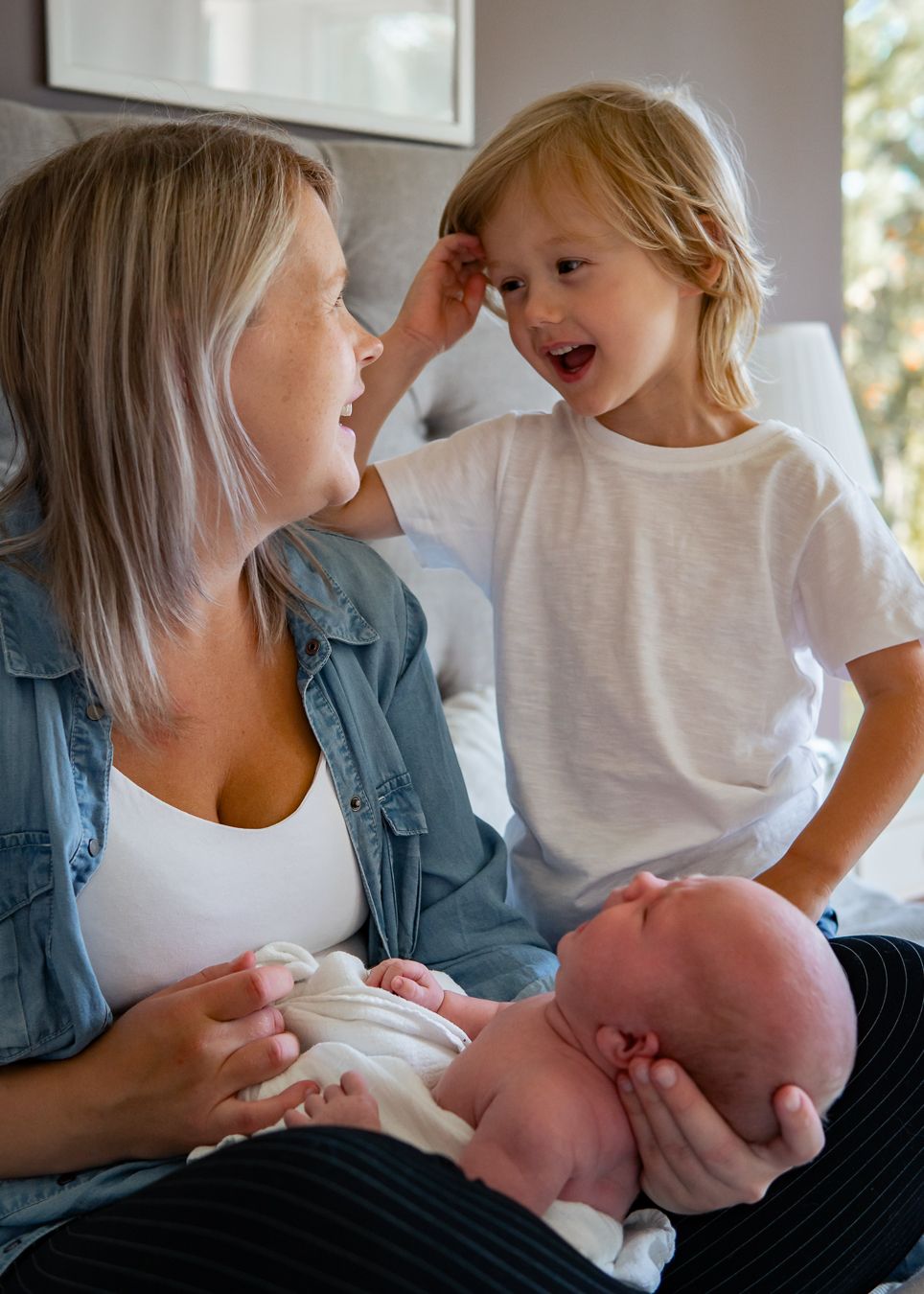 Ett mysigt familjeögonblick med skratt och samhörighet mellan mamma, son och bebis i sovrumsmiljö. Fotograf, Nyföddfotograf, Umeå
