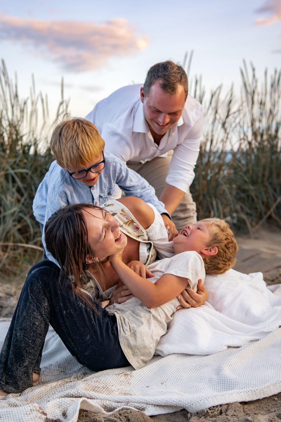 En familj leker och skrattar tillsammans på en filt på sandstrand i solnedgången med högt gräs i bakgrunden. Fotograf, Familjefotograf, Umeå