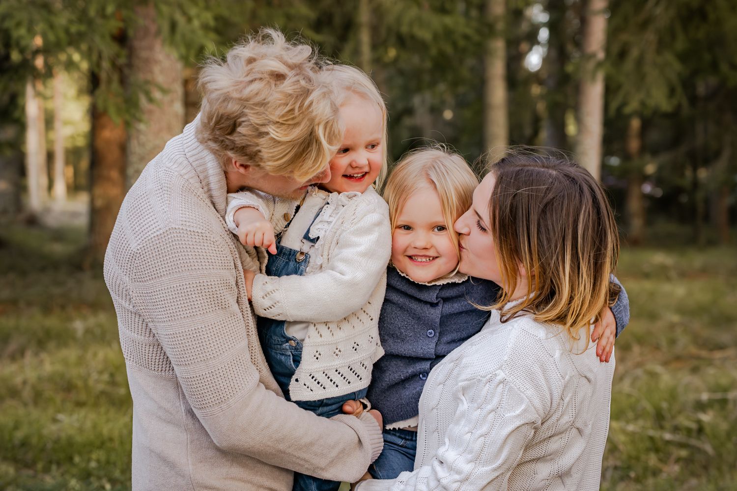 Kärleksfull familjebild där föräldrarna har barnen i famnen och pussar dem på kinderna med skogen som bakgrund. Familjefotograf, Umeå